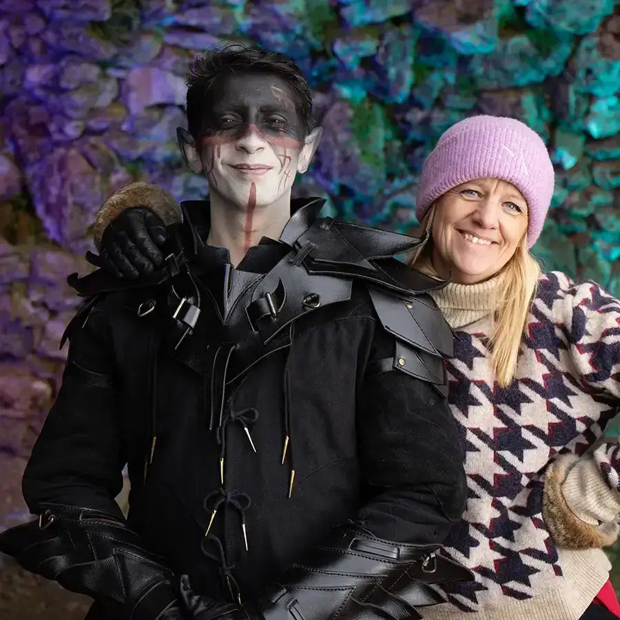 A person in dark fantasy costume with face paint poses with a smiling friend in cozy attire against a colorful stone backdrop.