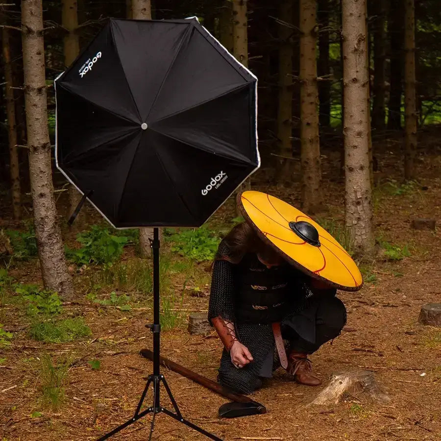 A photographer adjusting lighting equipment in a forest setting with a black umbrella softbox and an orange reflector.