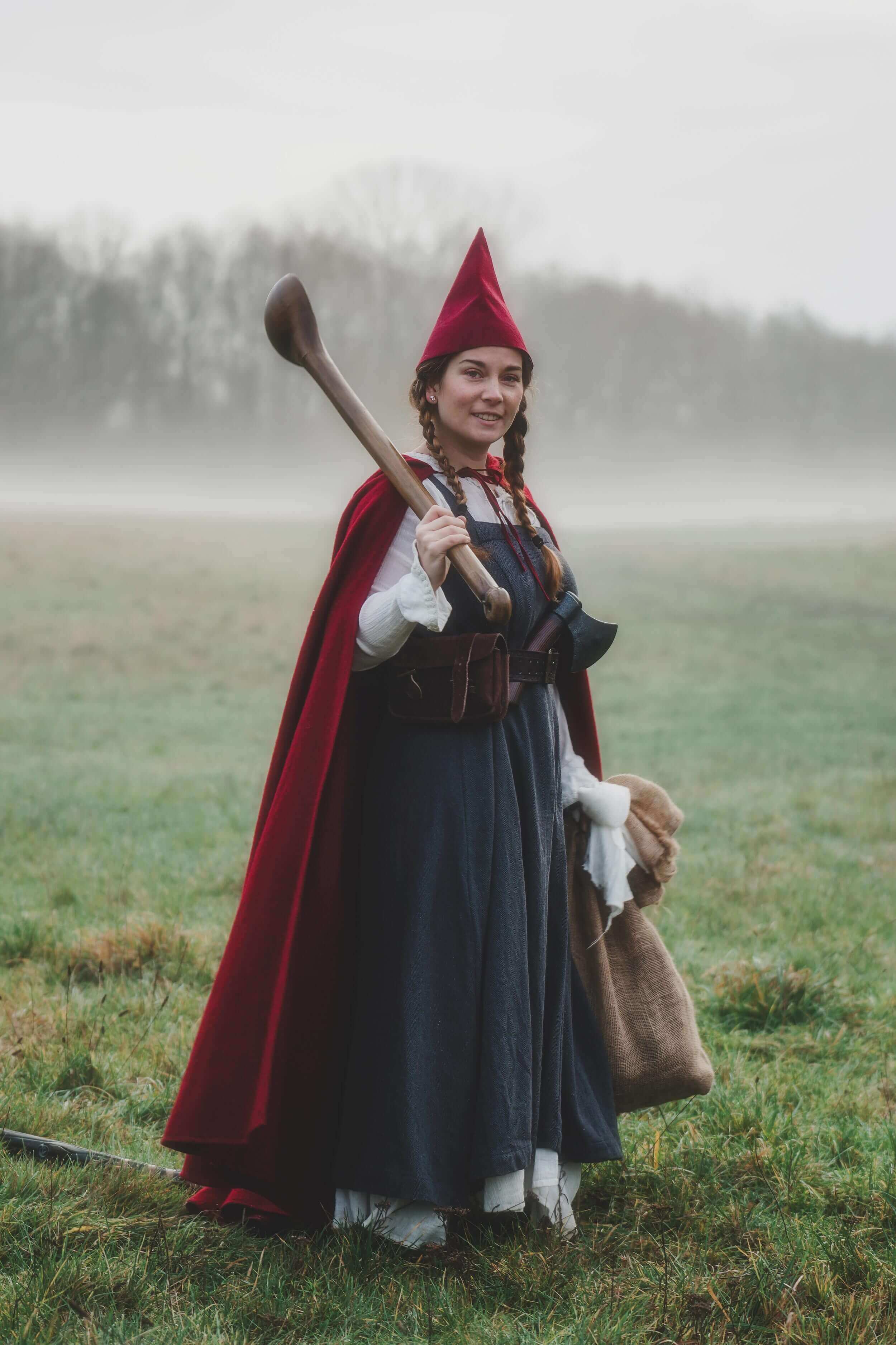 Woman in a red cloak and hat holding a wooden staff, standing in a misty field.