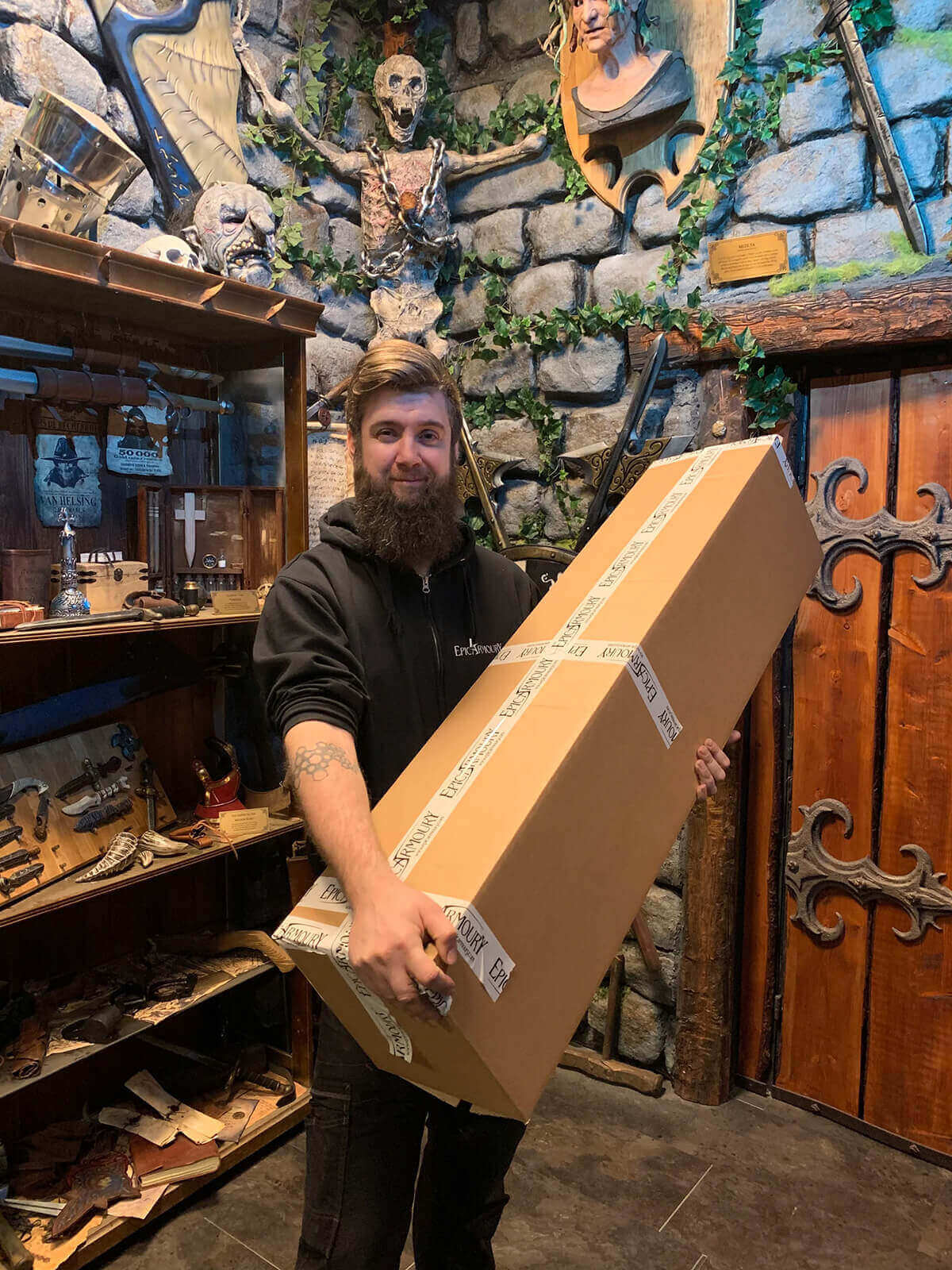 Man holding a large cardboard box in a rustic interior setting with stone walls and wooden furniture.