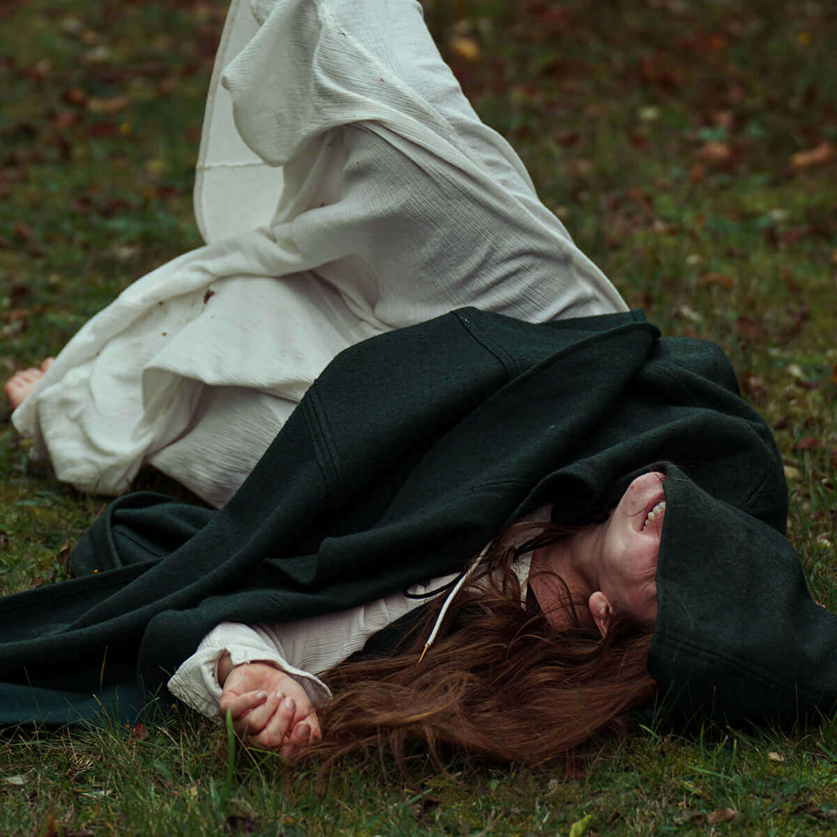 Woman with long red hair in a dark cloak, lying on grass, creating a dramatic and emotional scene.