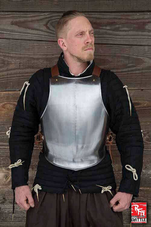 Man wearing historical armor with a metallic breastplate, standing against a wooden background.