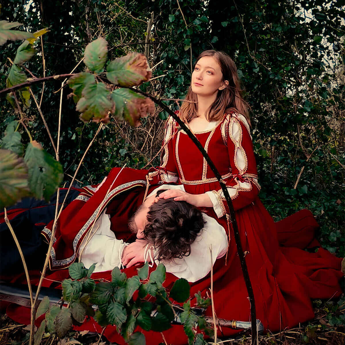 A woman in a red medieval dress sits beside a man reclining on the ground, surrounded by greenery.