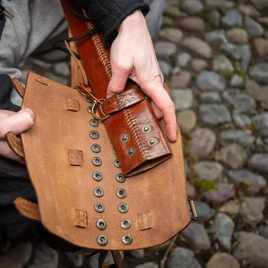 Person holding a rustic leather pouch with metal fasteners, set against a cobblestone background.