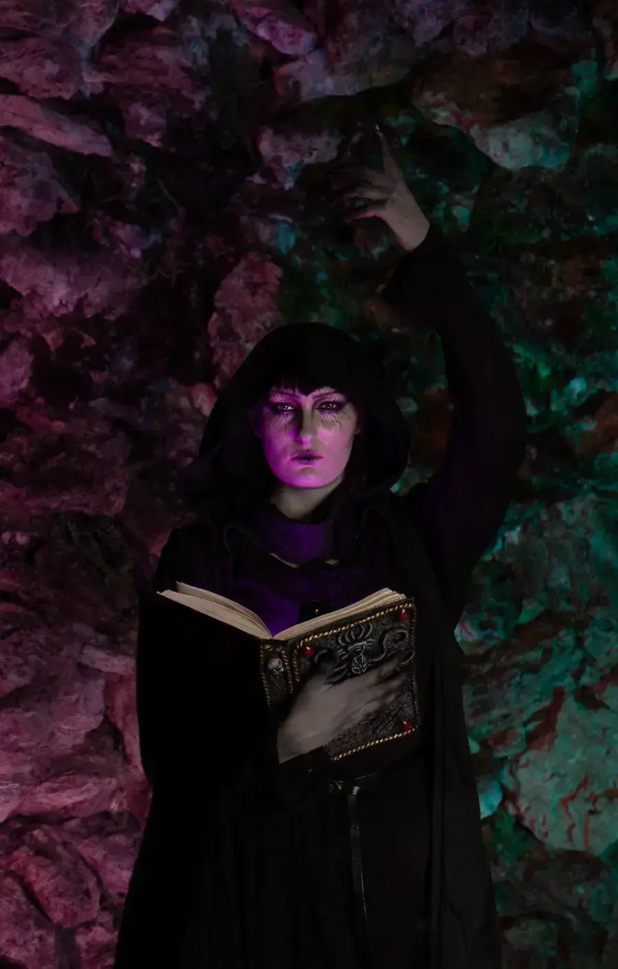 Mystical figure in dark cloak casting a spell while holding an ancient book against a colorful, rocky backdrop.