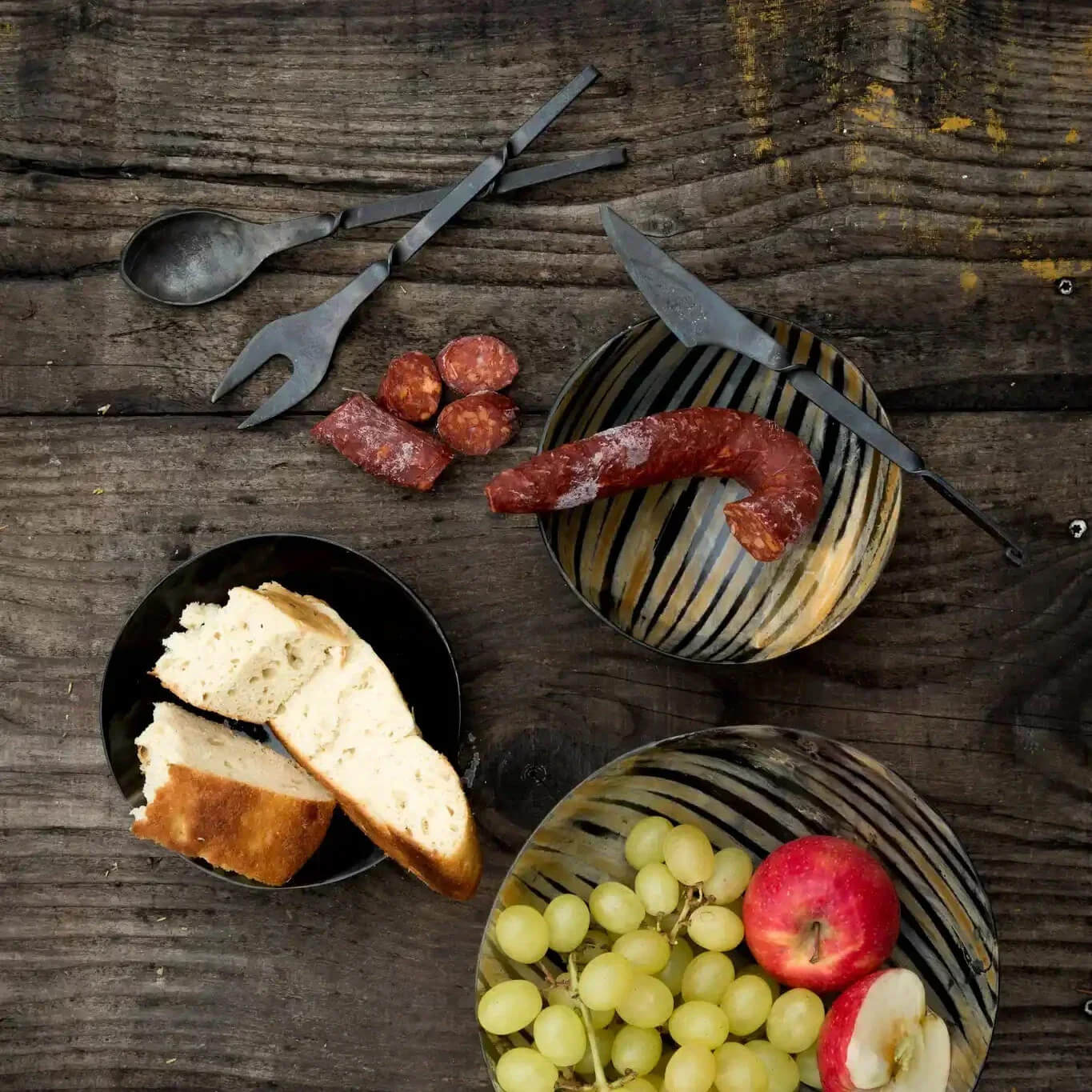 A styled flat-lay of rustic black metal serving utensils, including a spoon, fork, knife, and platter, arranged on a dark wooden table with food props like sliced bread, sausage, grapes, and an apple.
