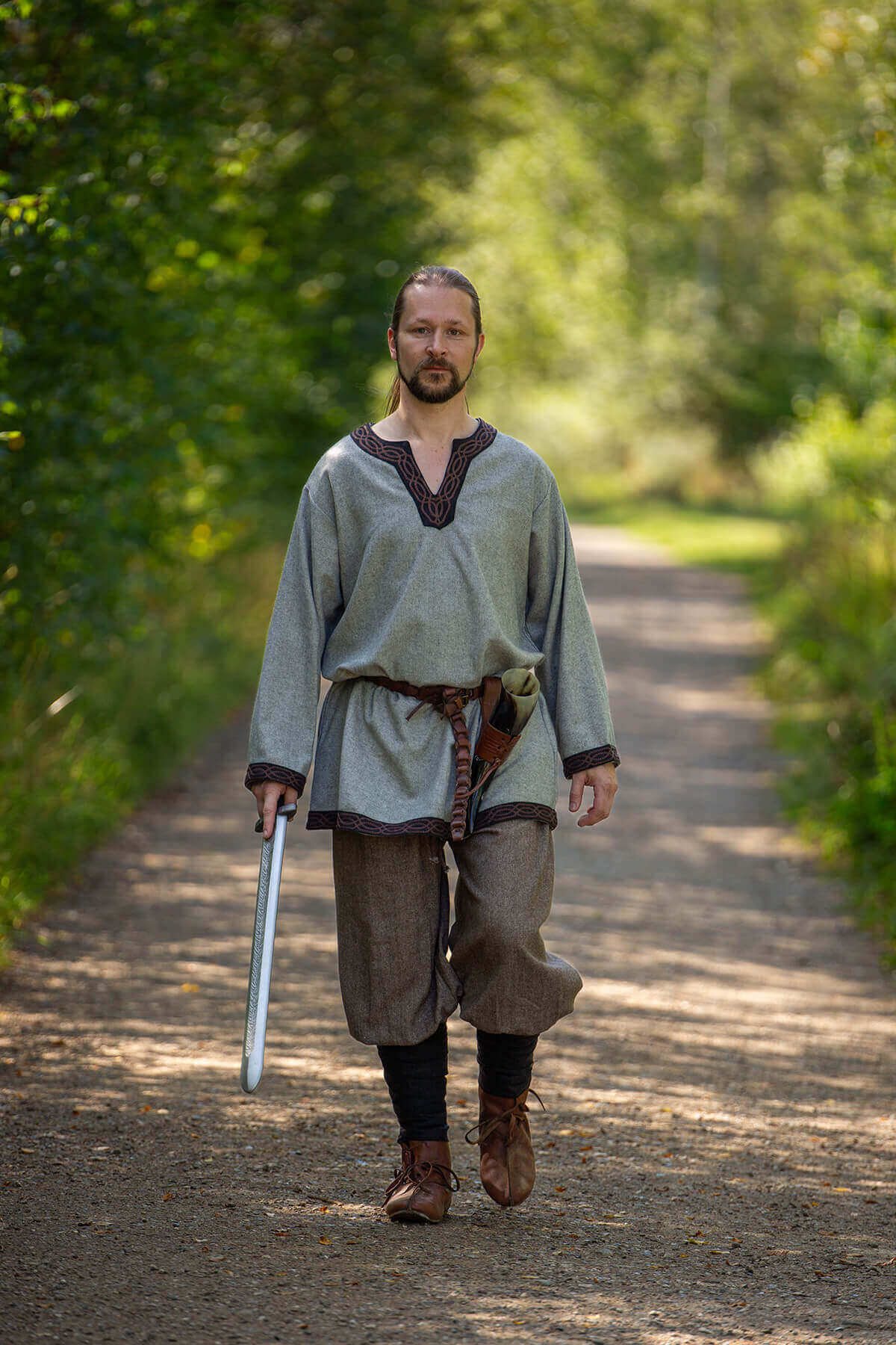 Man in historical attire holding a sword on a path surrounded by trees