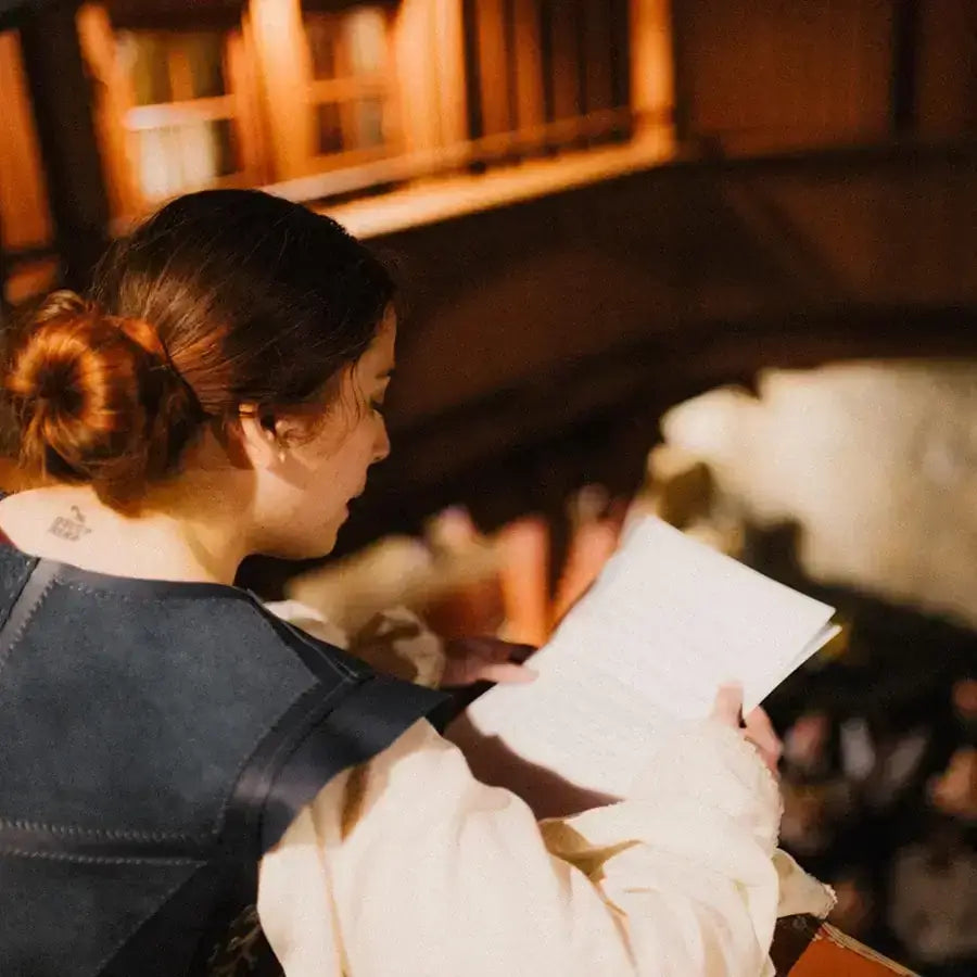 Woman in historical dress reading a book indoors with wooden architecture in the background.