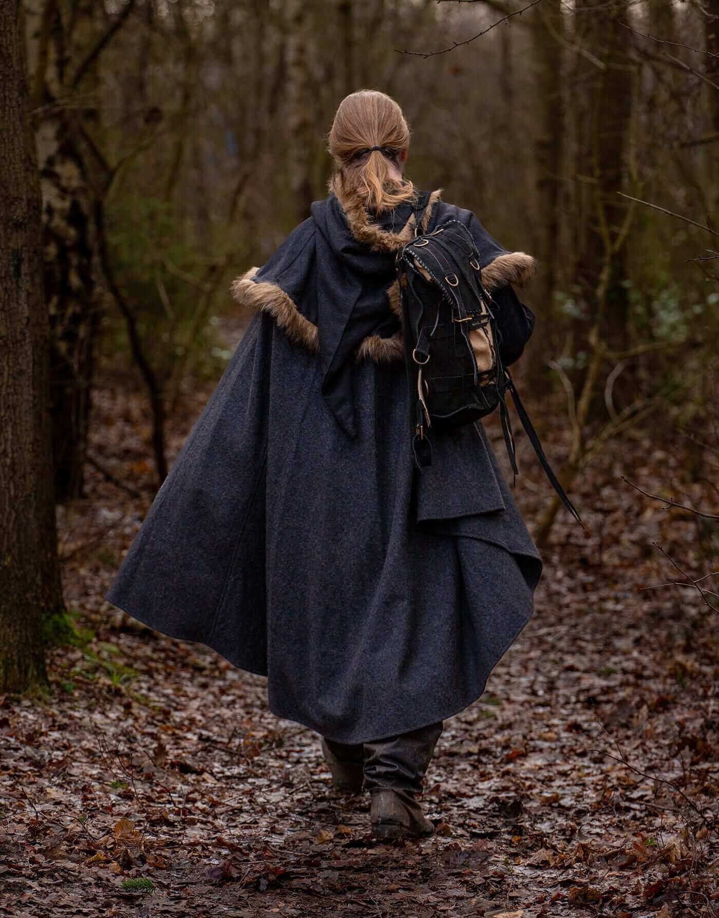 Woman in a dark cape walking through a wooded path with a backpack, surrounded by autumn leaves.