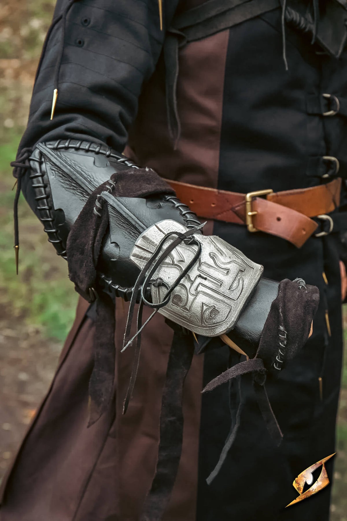 A close-up of a person wearing a detailed black leather and fabric arm guard with a silver engraved metal plate, dressed in a dark brown and black costume outdoors.