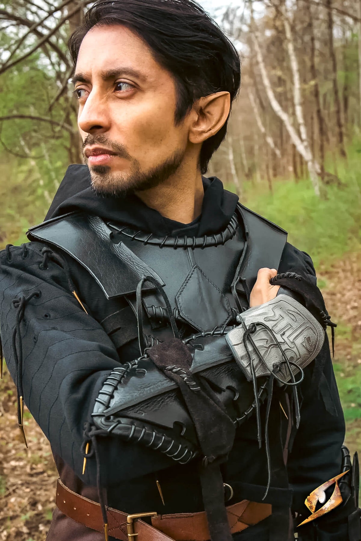 A man wearing a detailed fantasy costume with a black hooded tunic, dark leather-look armor plates, and a gray laced gauntlet, standing in a wooded outdoor setting.