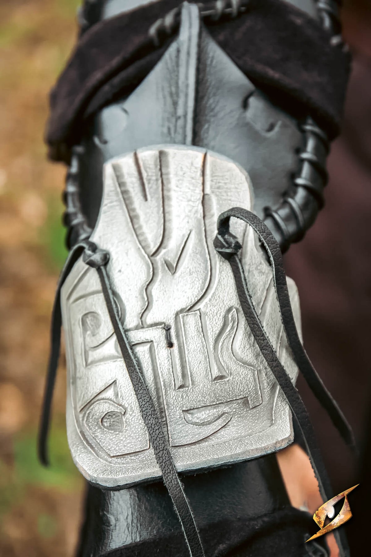 A close-up of a black leather bracer or gauntlet with a large engraved silver metal plate and black laces, shown outdoors against a blurred natural background.