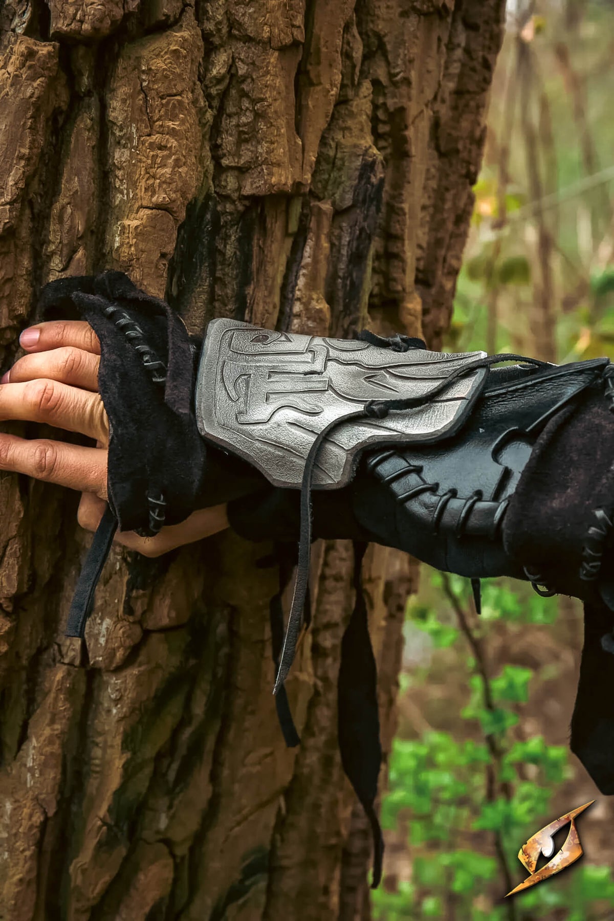 A close-up of a person's hand resting against a textured tree trunk, wearing a black leather wrist guard with a silver metallic forearm plate and black gloves.