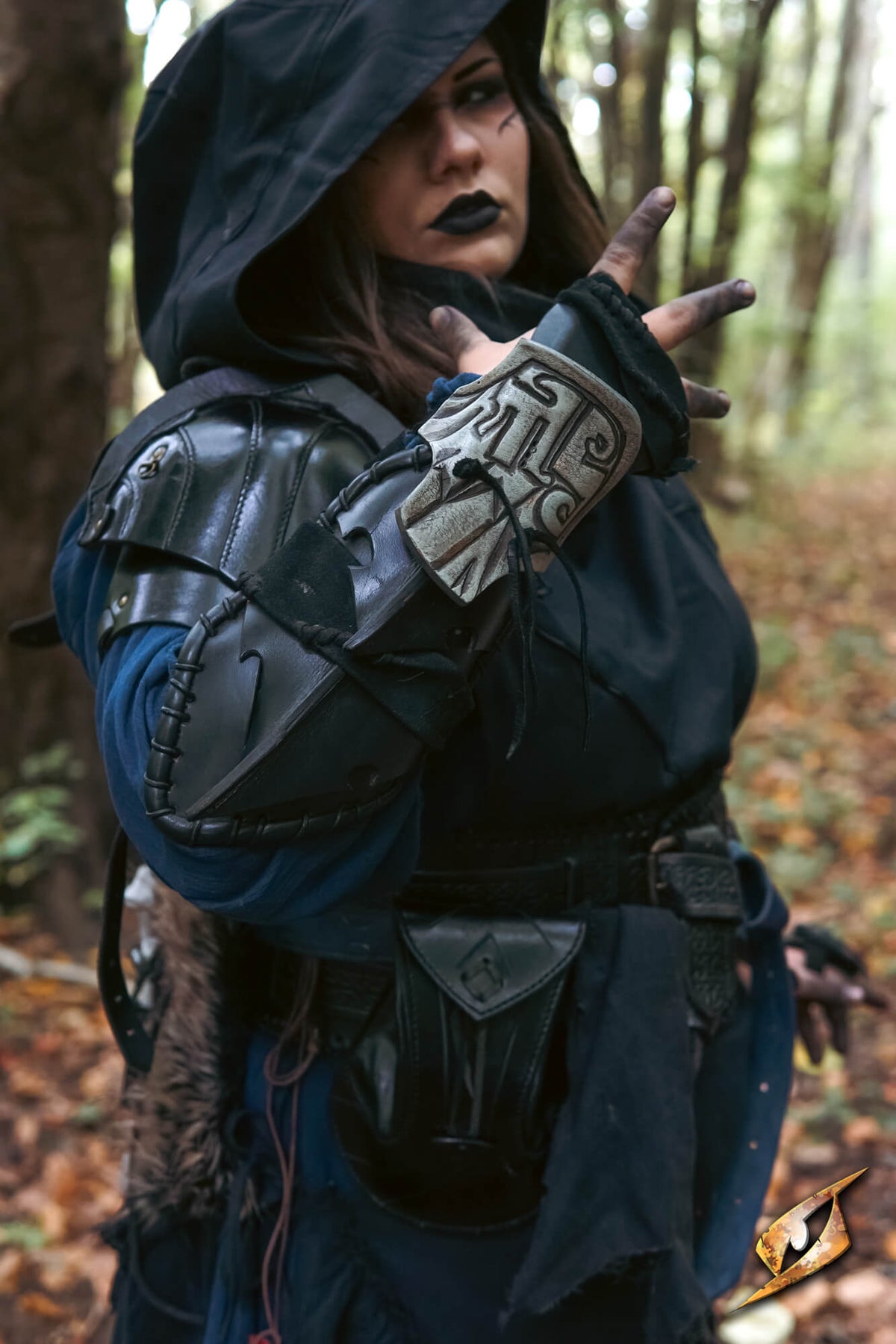 A person in a fantasy warrior costume stands in a forest, wearing a black hooded tunic, dark leather-like armor pieces, a blue underlayer, and a white decorative wrist cuff.