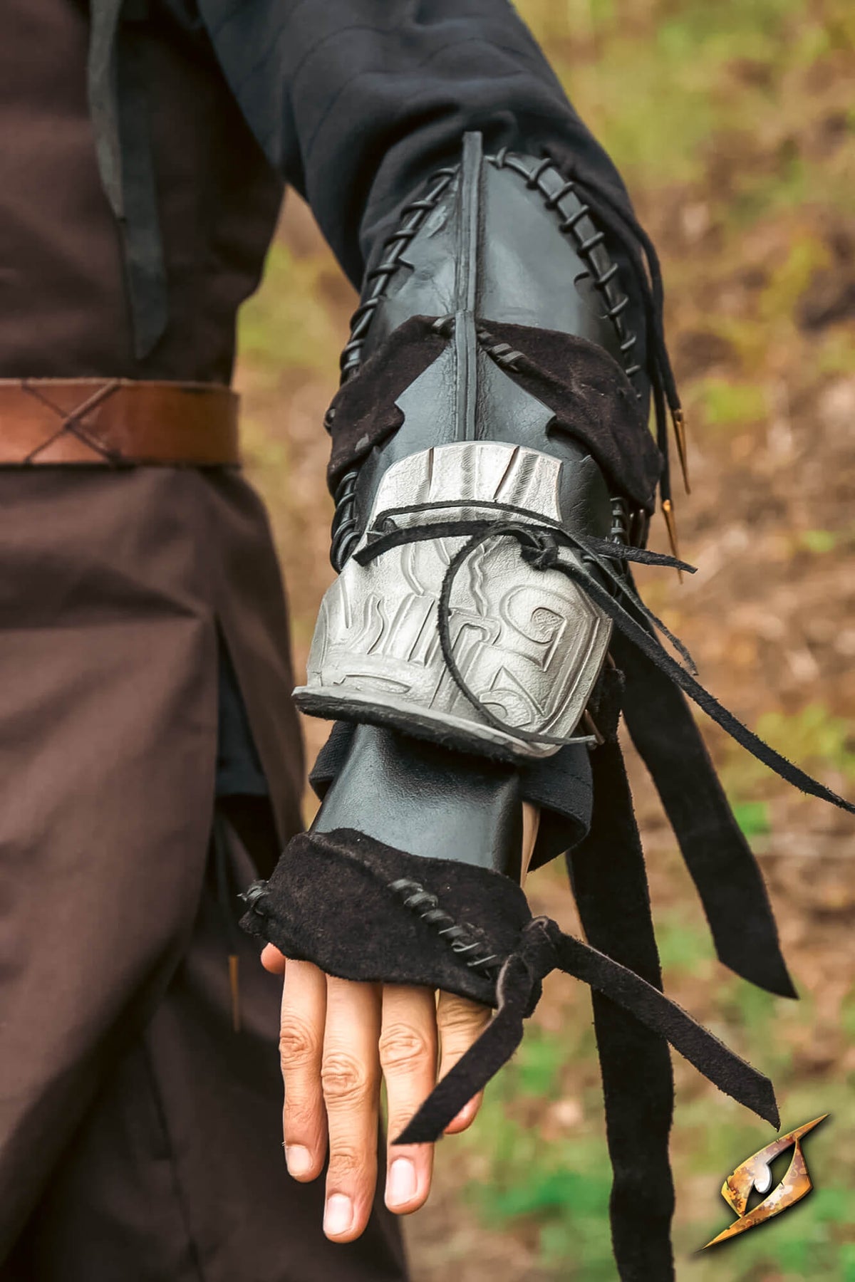A close-up of a person's forearm wearing a dark brown and black fantasy-style leather bracer with a carved bone-like guard and black laces, set against a blurred outdoor background.