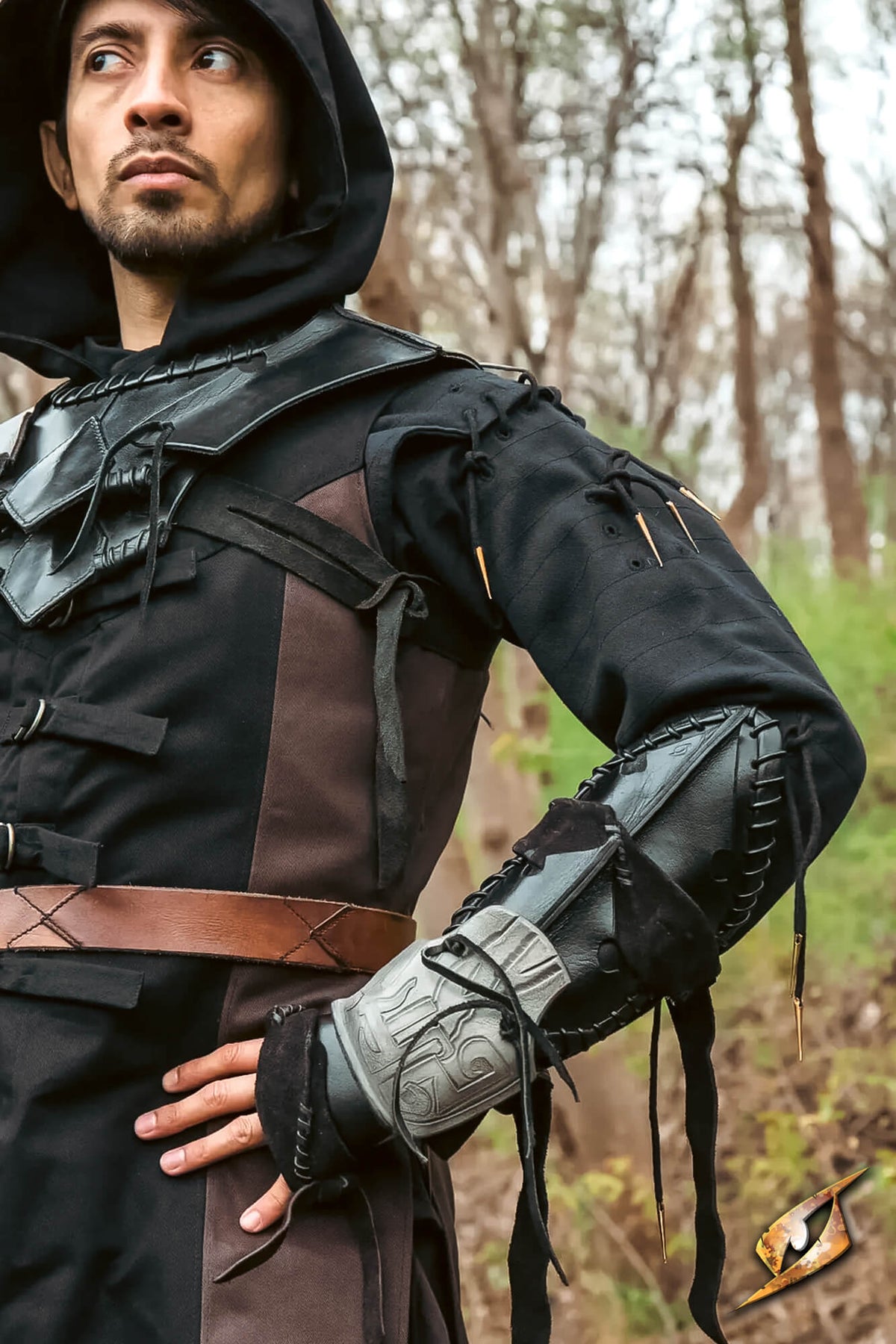 A man wearing a detailed fantasy costume, featuring a black hooded tunic with brown leather-like panels, a brown belt, and a black-and-silver armored forearm guard, posed in a forest setting.