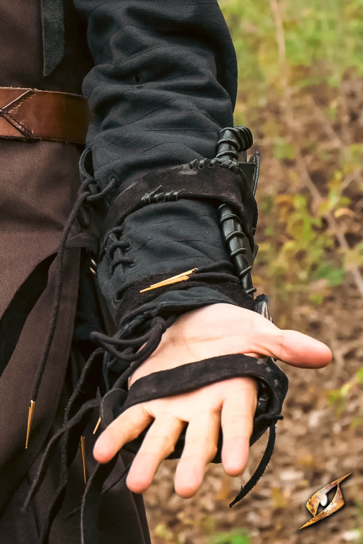 A close-up of a person's hand wearing a black leather pirate-style wrist guard with laces, brass pins, and a dark strap, set against an outdoor blurred background.