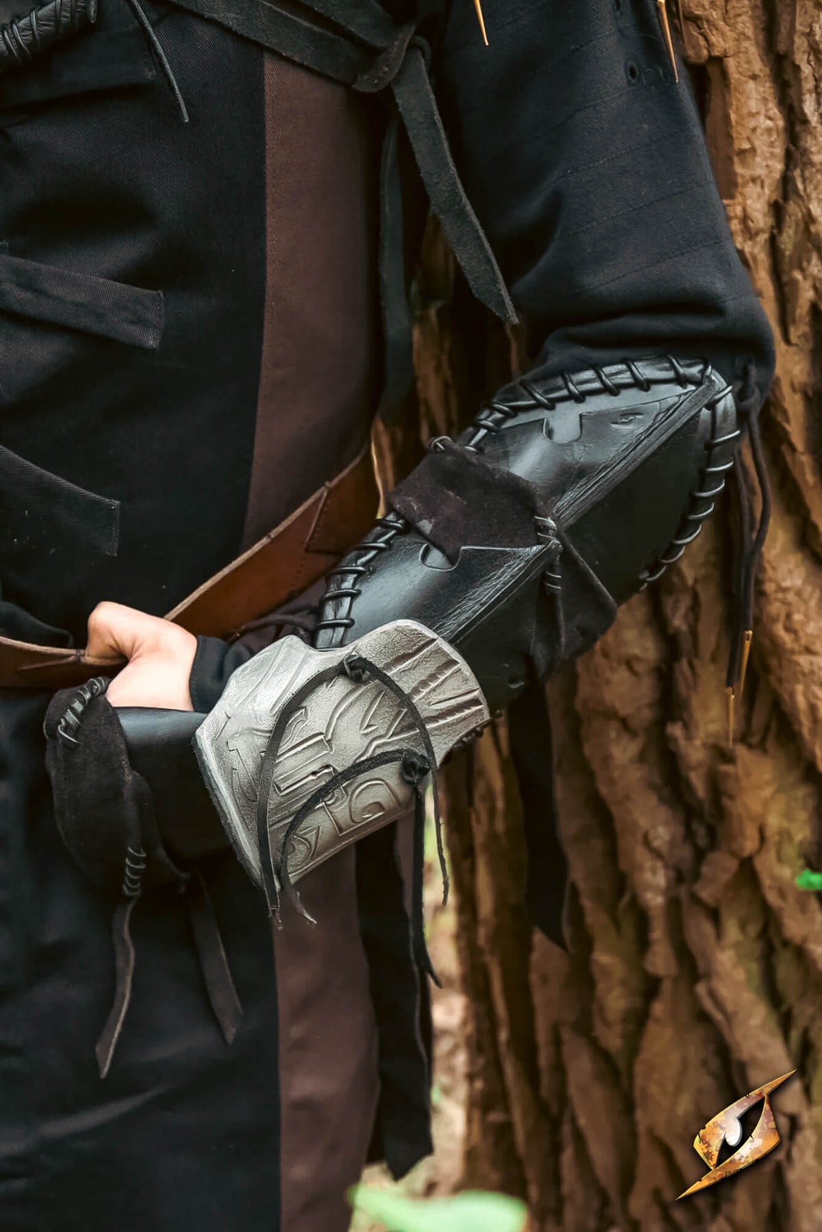 A close-up of a person wearing a black leather and faux-leather arm guard with stitched detailing and a gray sculpted forearm plate, posed in front of a textured tree trunk.