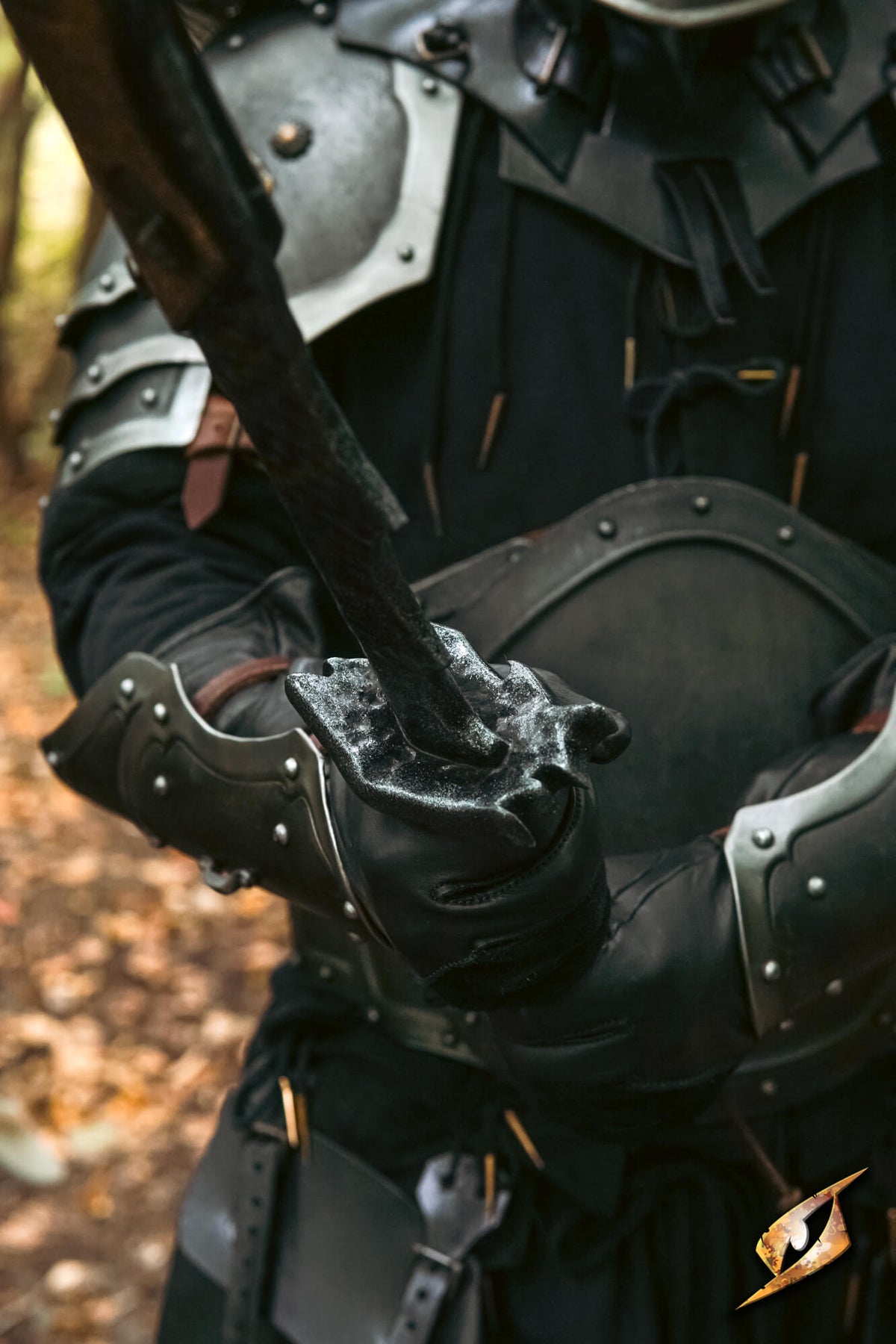 Close-up of a person wearing dark medieval-style armor and black gloves, holding a sword in an outdoor wooded setting.