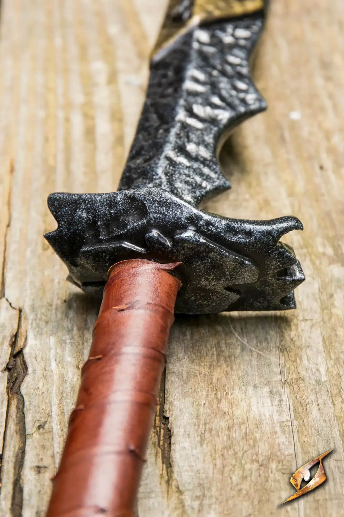 A close-up of a fantasy sword with a textured dark gray blade, a dragon-head pommel, and a brown leather-wrapped grip, resting on a rustic wooden surface.