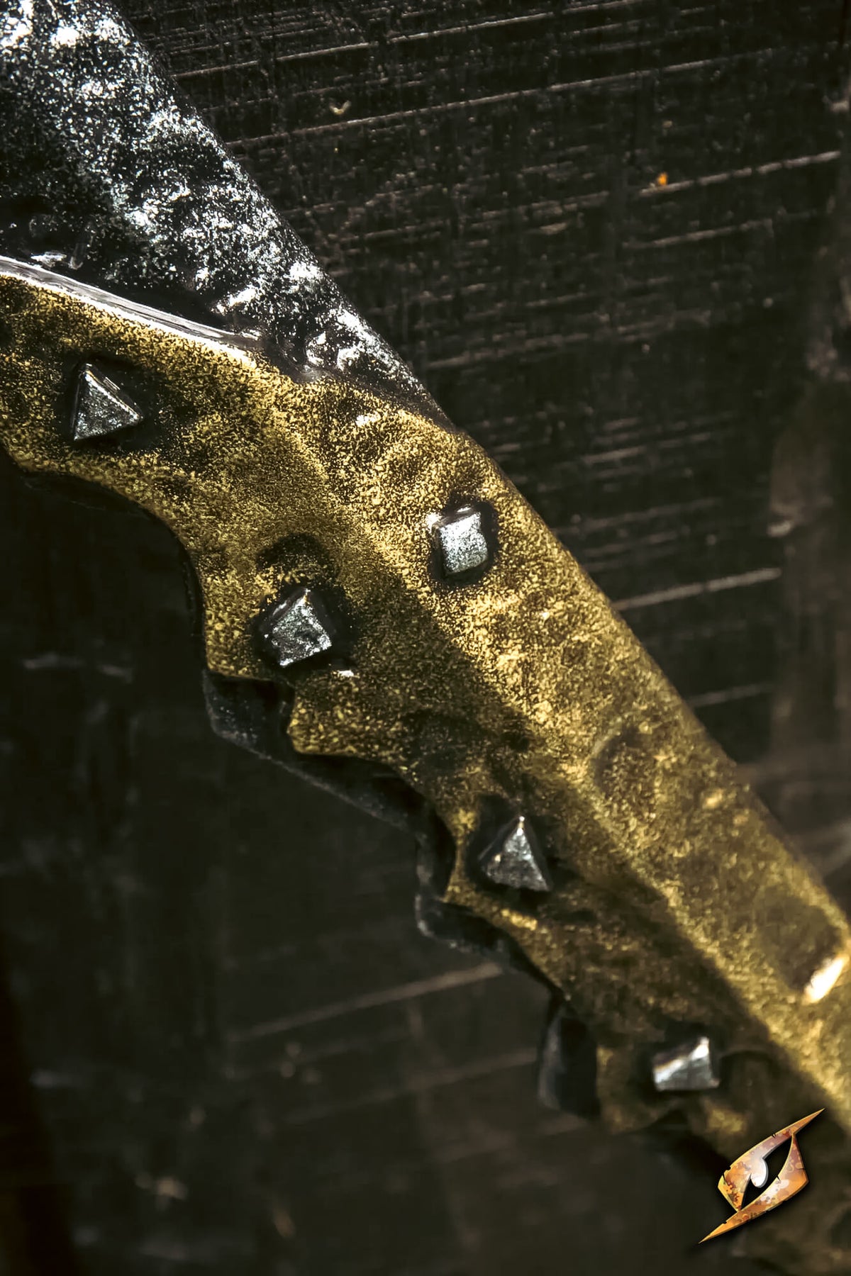 A close-up of a fantasy blade with a textured gold and black finish, featuring raised triangular and square metallic studs along the edge, set against a dark textured background.