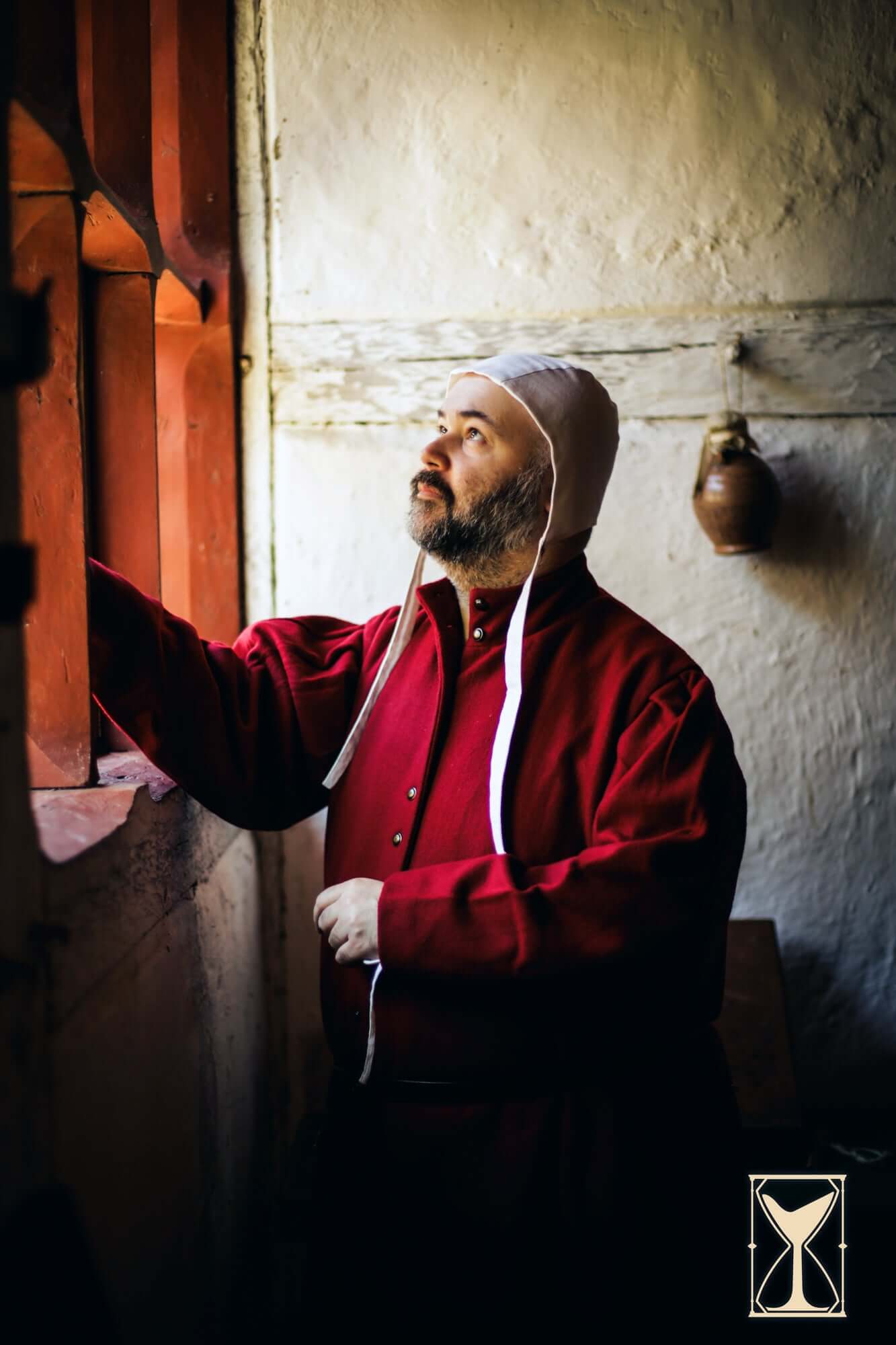 Man wearing a red garment and hat looking out a window, showcasing the Surcoat Lorenzo style.