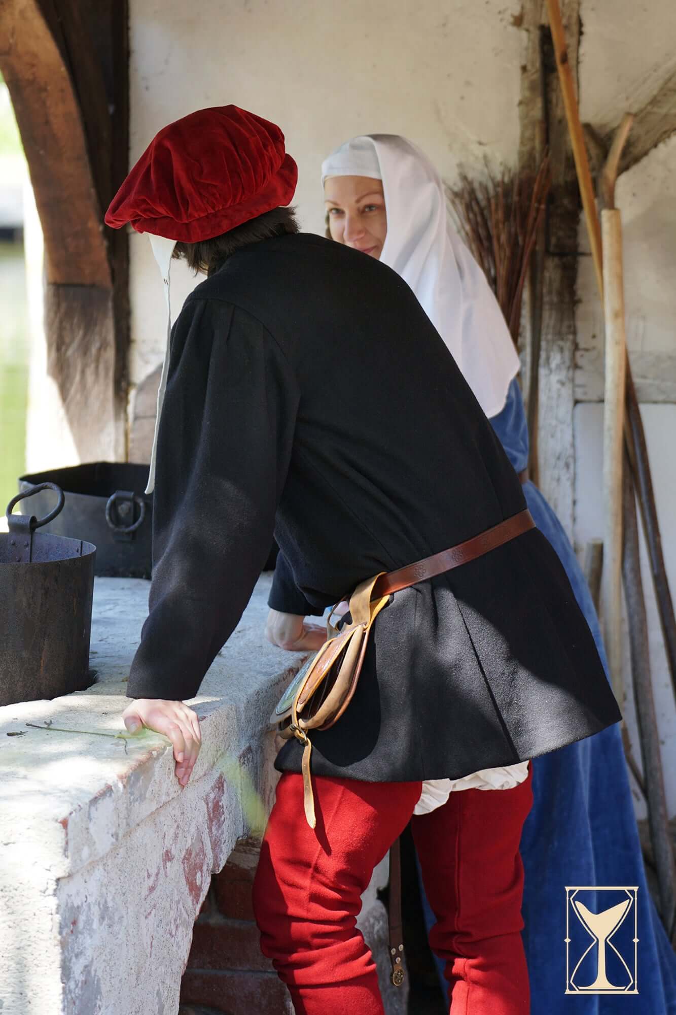Two figures in historical attire conversing near a rustic table, showcasing traditional 14th and 15th-century clothing.