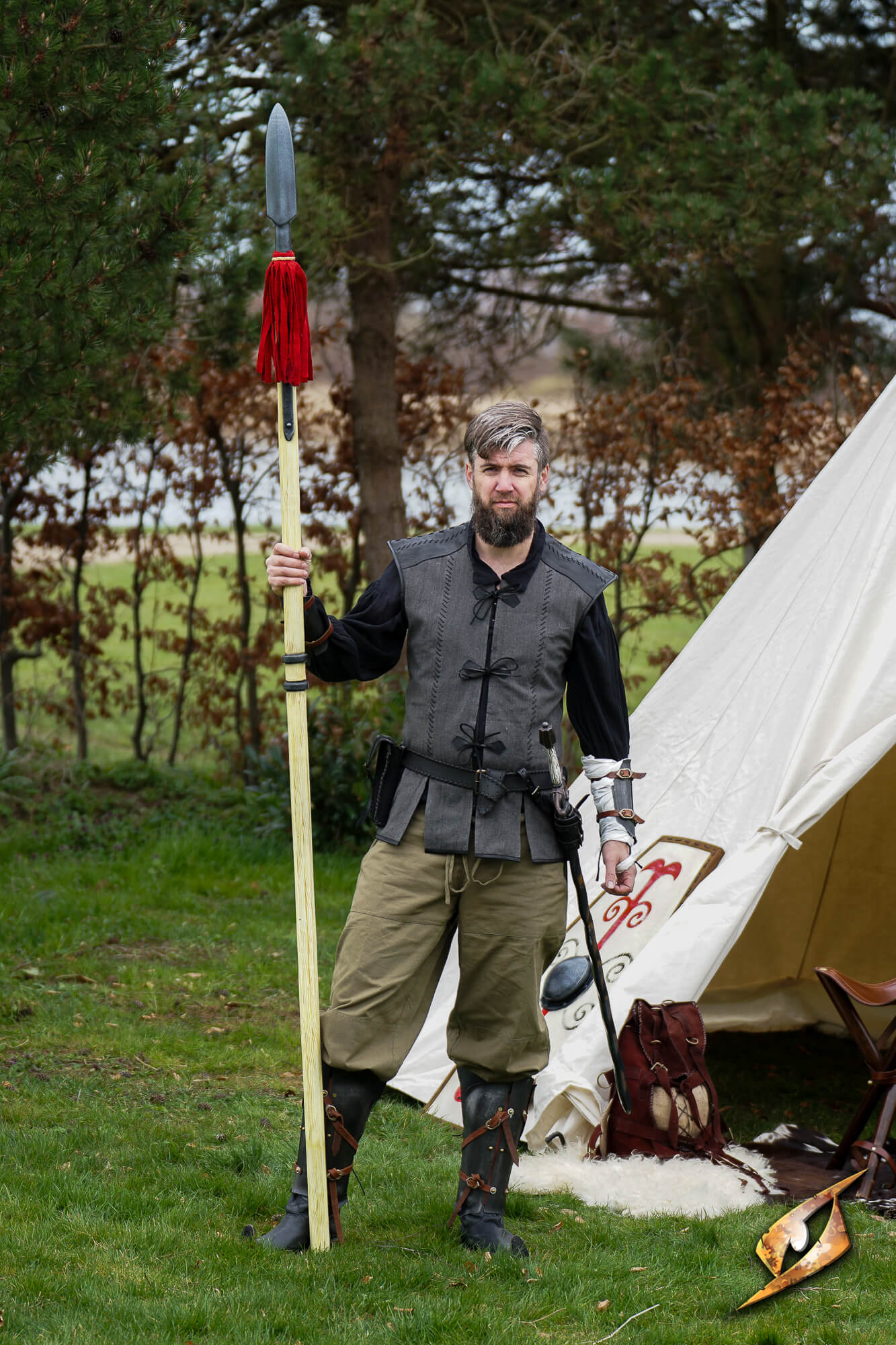 Man in medieval attire holding a spear next to a tent outdoors