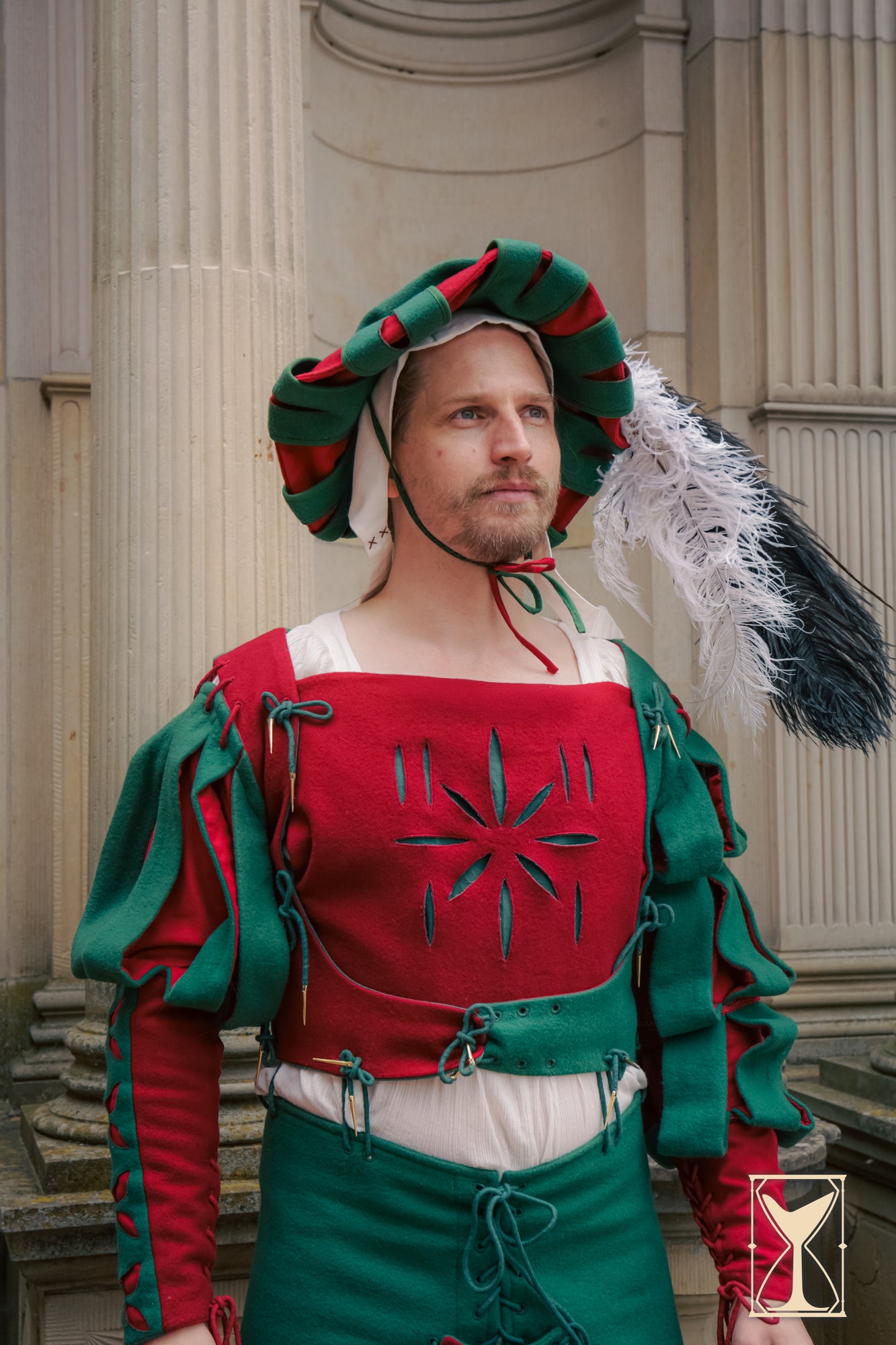 Person in a red and green costume with feathered headdress standing in front of classical architecture.