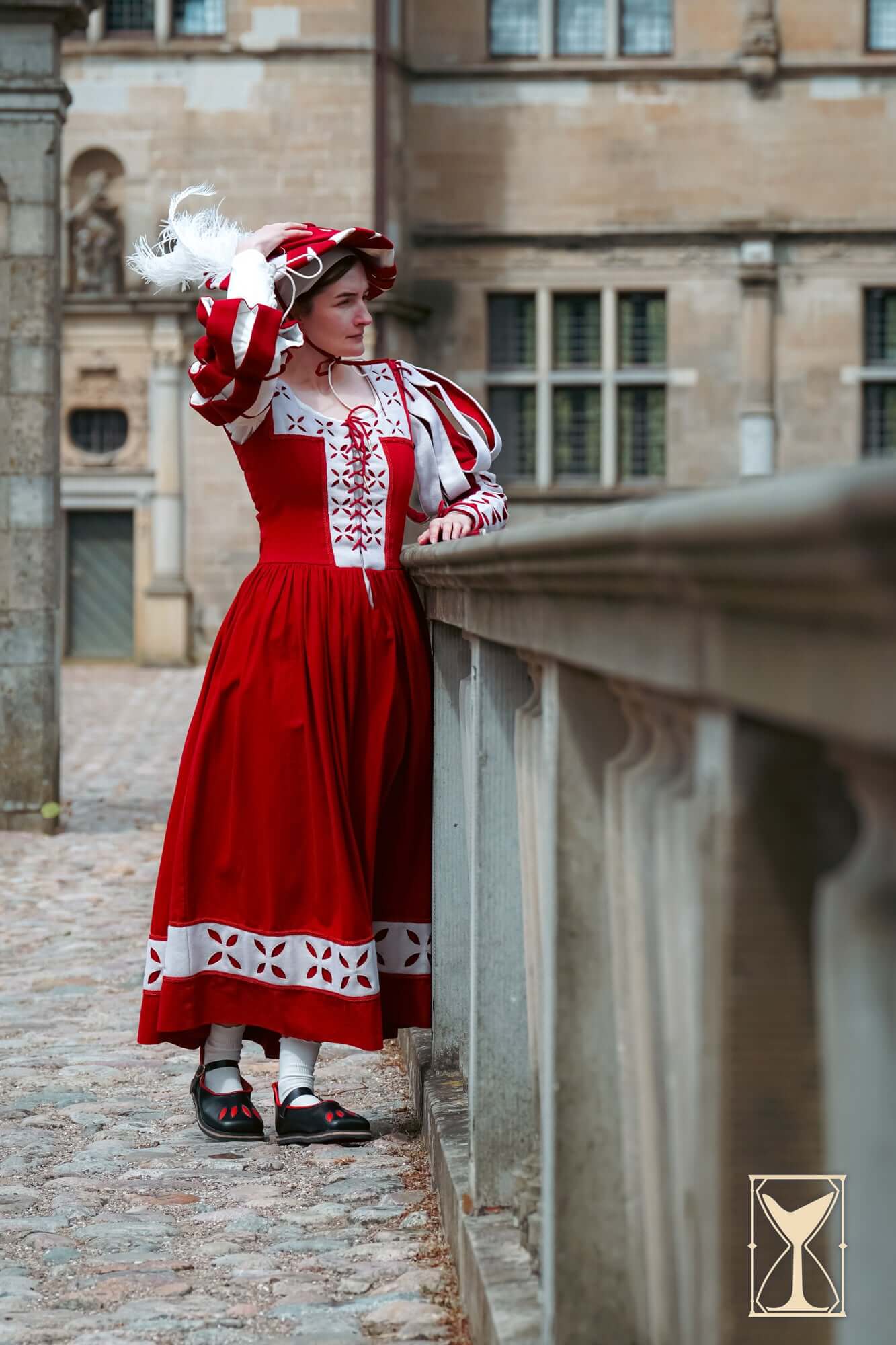 Woman in a traditional Renaissance costume featuring a red dress and white accents, standing by a stone railing.