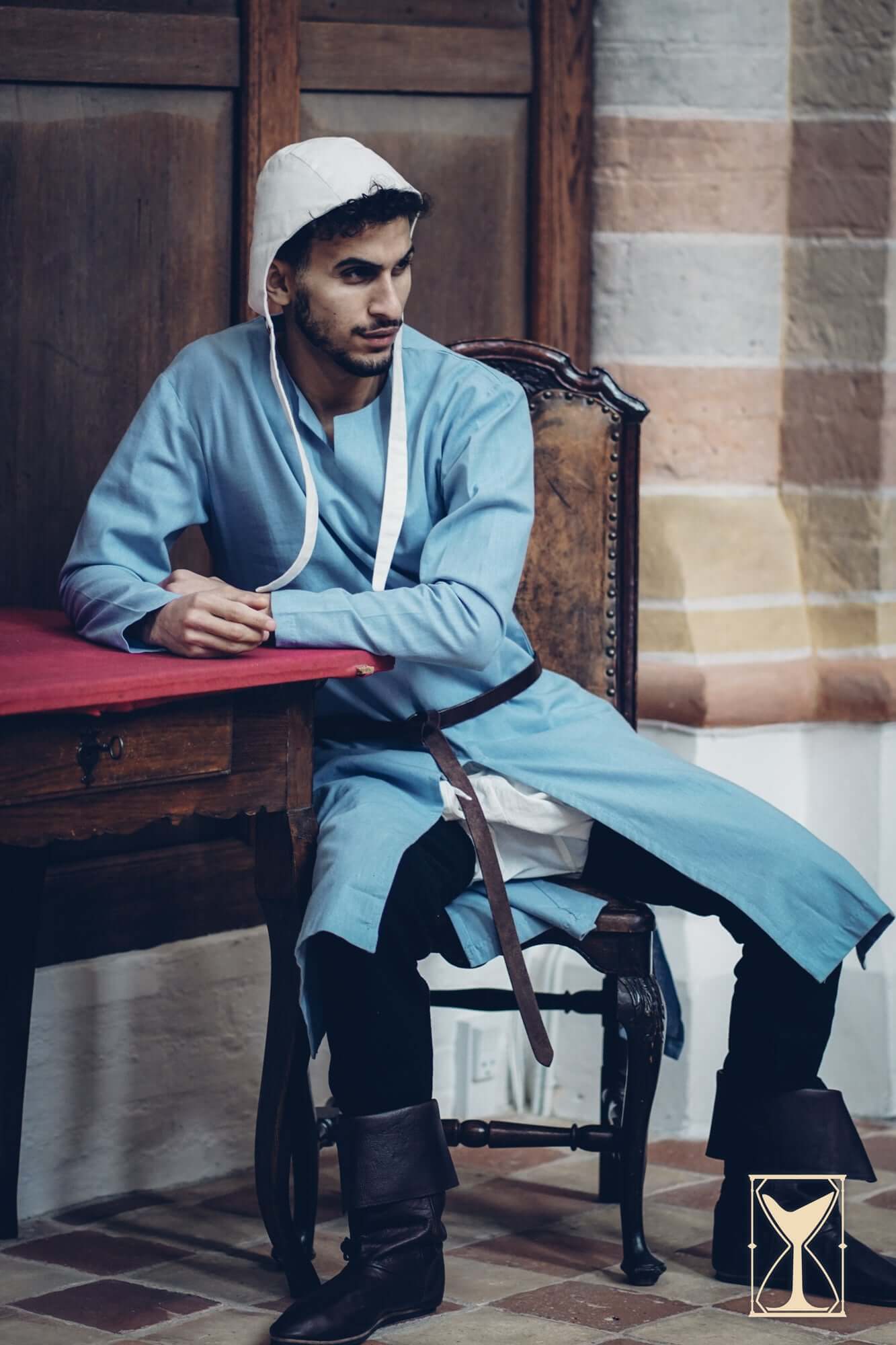 Man in a blue Medieval Riding Tunic seated at a wooden table, showcasing historical medieval attire.