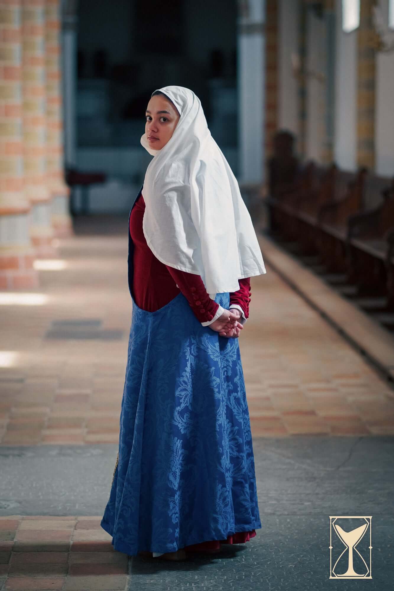 Pellote - Medieval Dress: A model wearing a sleeveless surcoat with a white hood and blue skirt in a historical setting.