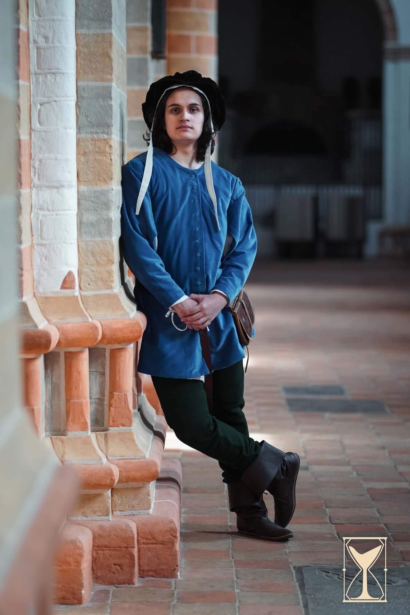 Person wearing a blue tunic and hat in medieval setting, showcasing the Velvet Surcoat as a stylish outer layer.