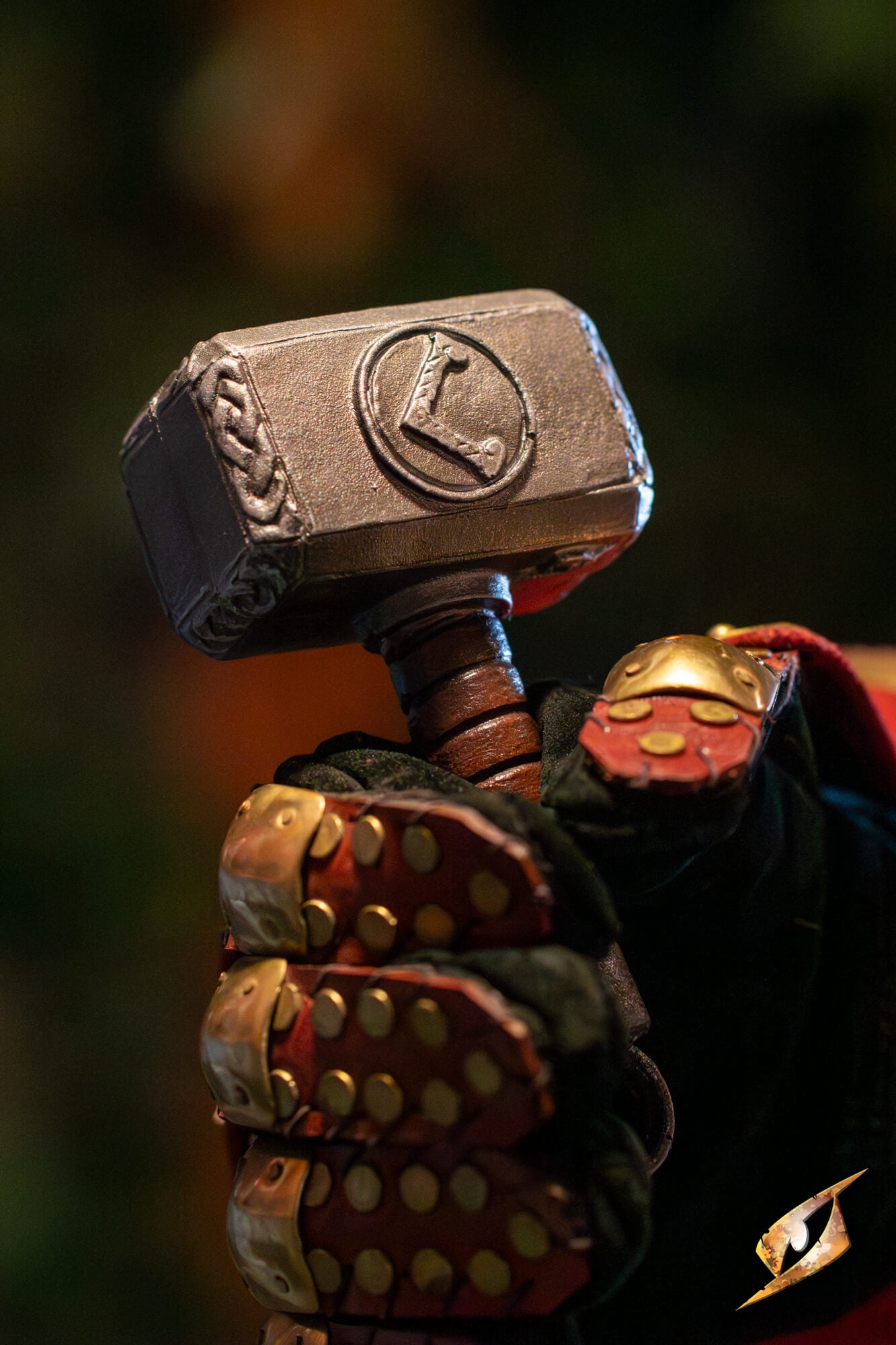 A close-up of a hand wearing a detailed Thor costume glove holding a replica of Mjolnir, with a blurred dark background.