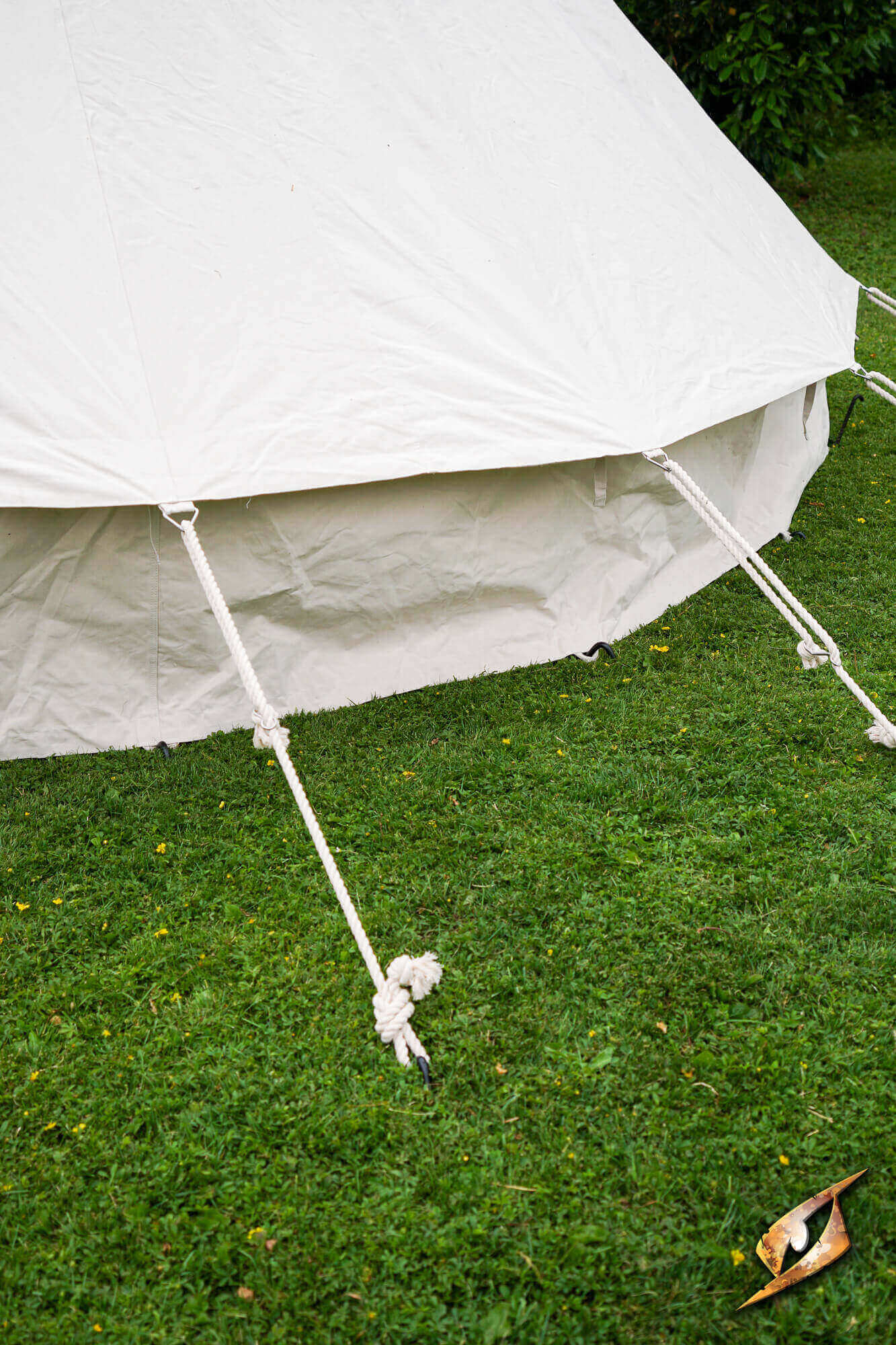 A close-up outdoor shot of a white canvas bell tent pitched on green grass, with tent ropes anchored into the lawn and a small dry leaf in the foreground.