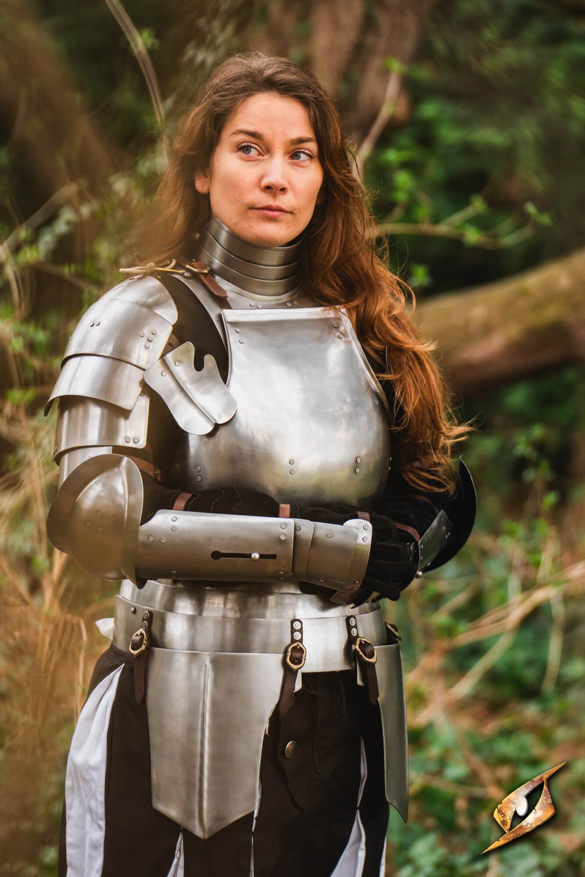 A woman wearing medieval-style steel plate armor stands outdoors in a forest setting. The armor includes a breastplate, gauntlets, and a chainmail neck cowl, with black and white fabric layers visible underneath.