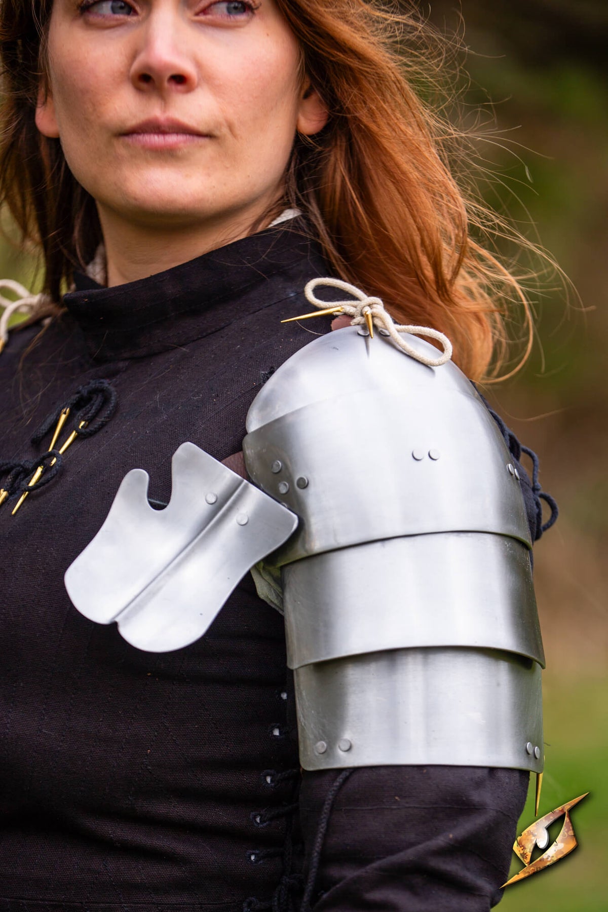A close-up of a woman wearing medieval-style steel plate armor on her shoulder, including a large pauldron and smaller shoulder guards over a dark tunic, with a blurred outdoor background.