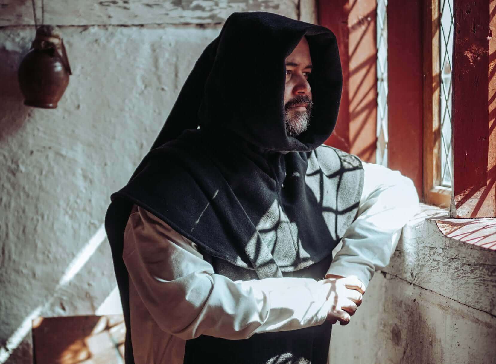 Man in a hooded cloak standing by a window, sunlight casting shadows on the wall.