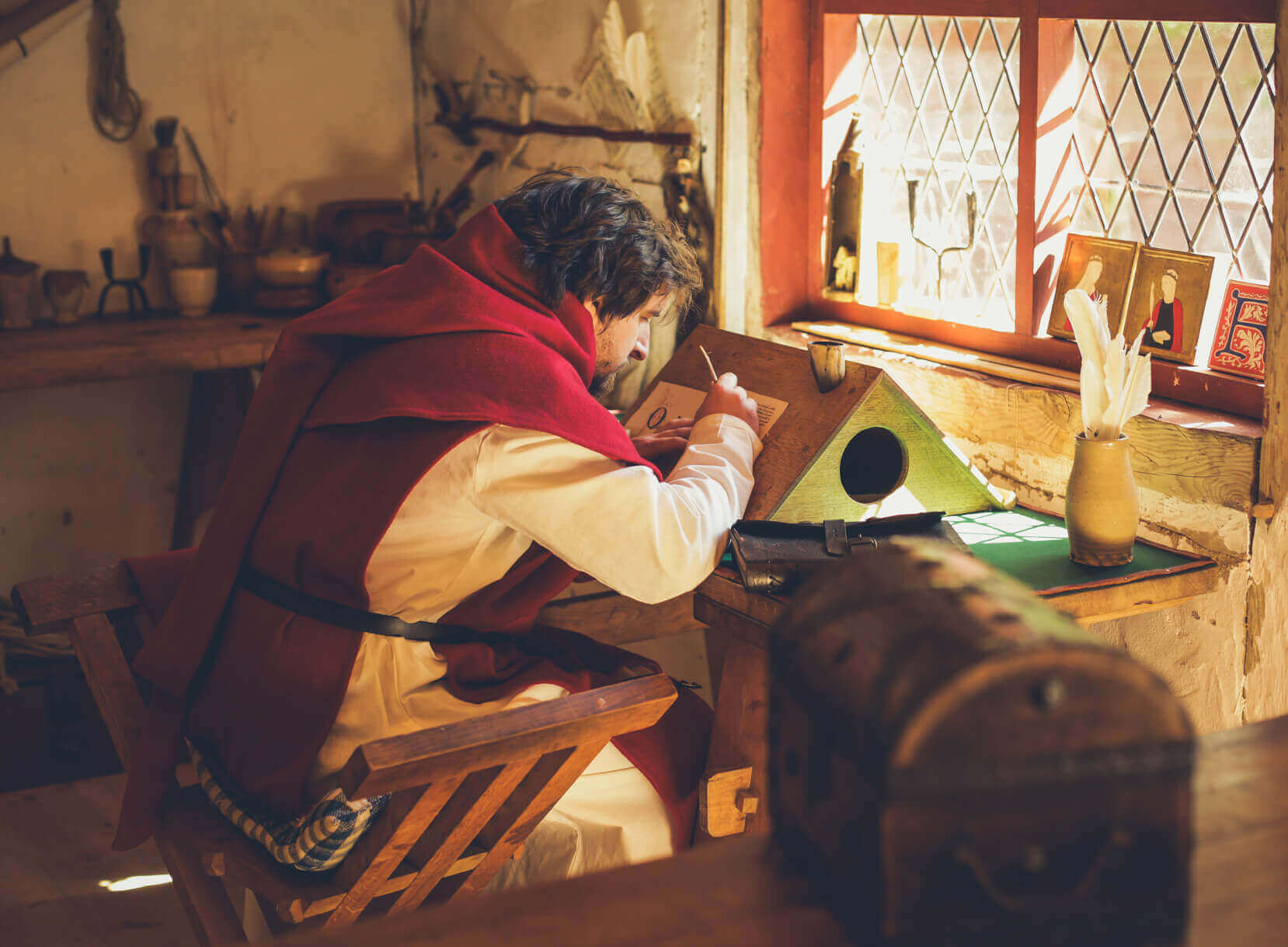 Person in historical clothing writing at a desk by a window, surrounded by vintage decor and cozy lighting.