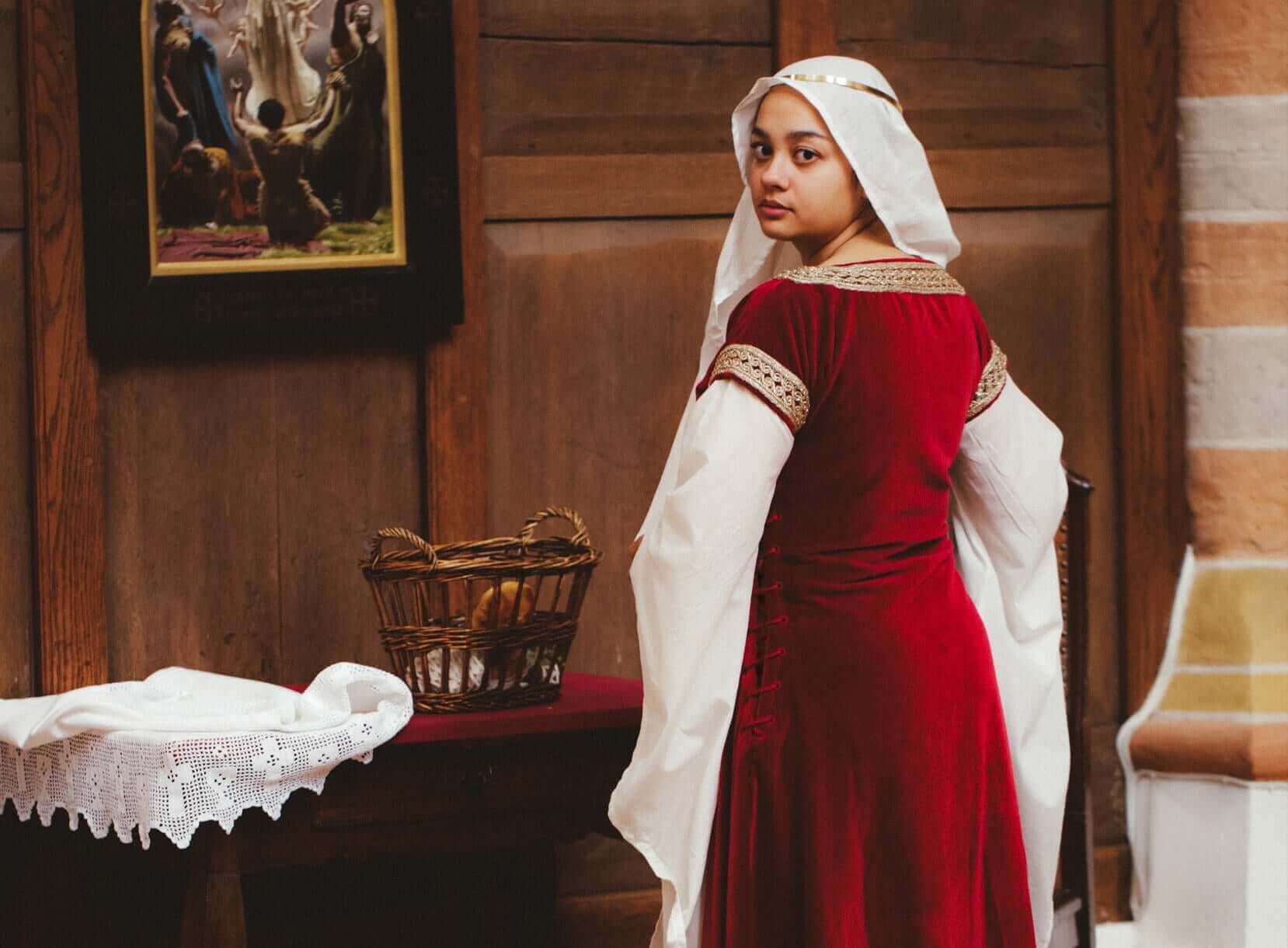 Person in a red dress with white sleeves standing in a room with wooden walls and a basket on a table.