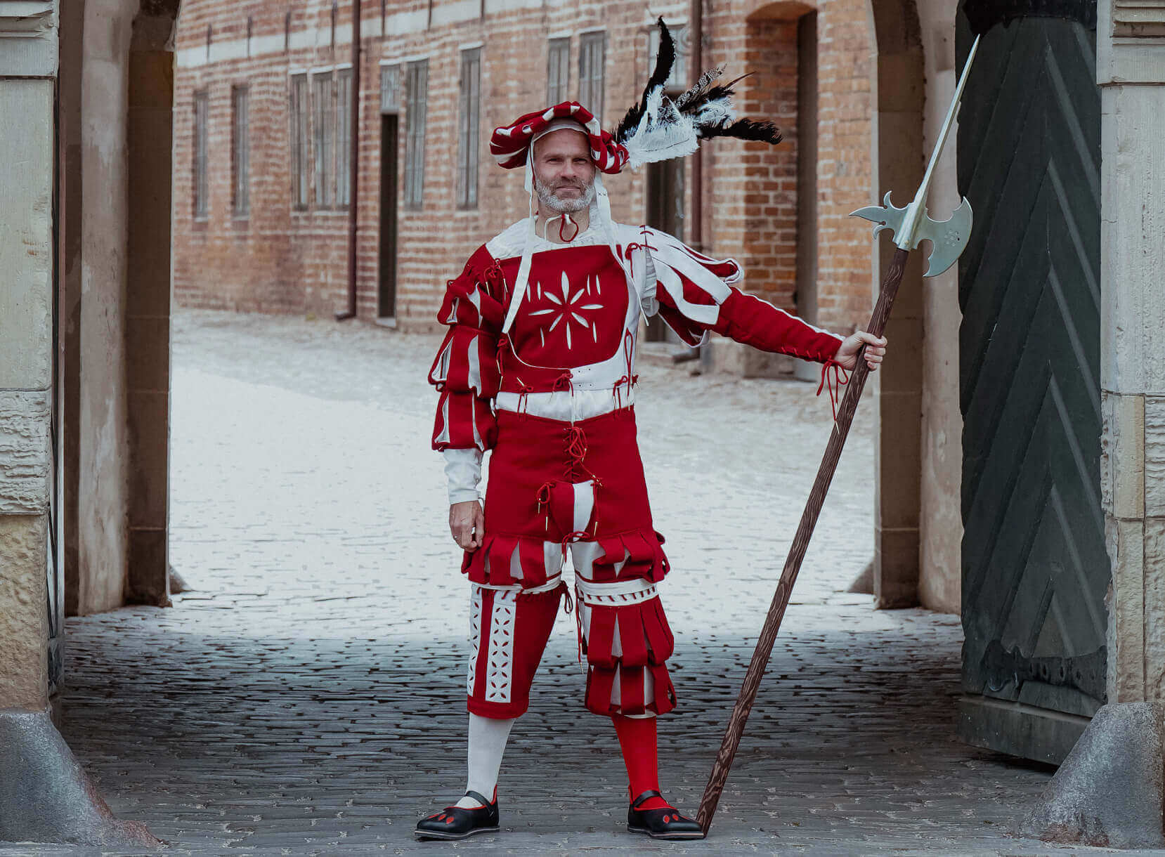 Historical performer in red costume and feathers stands with a spear in a medieval setting.