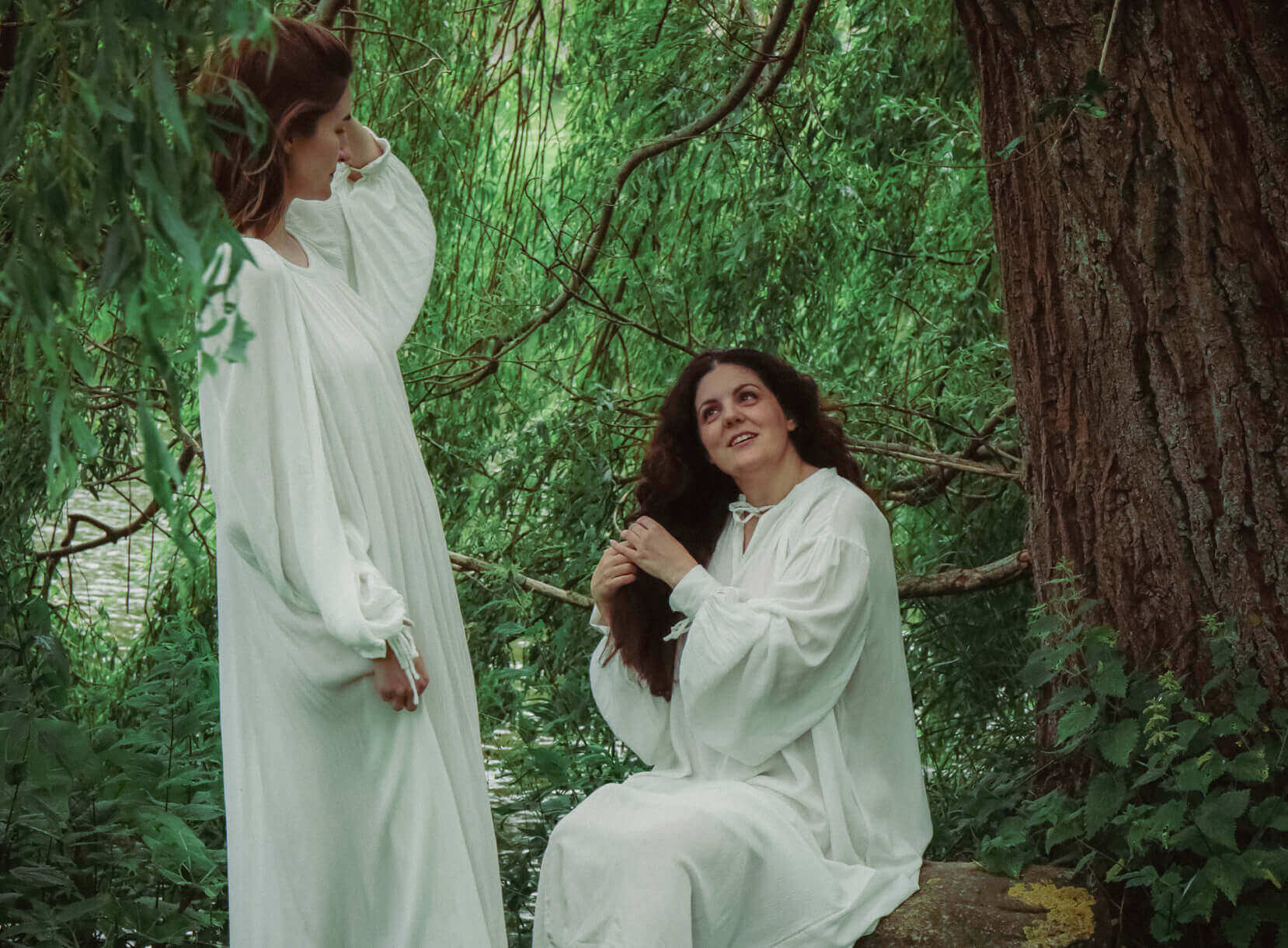 Two women in flowing white dresses, one braiding the other's hair in a lush green forest setting.