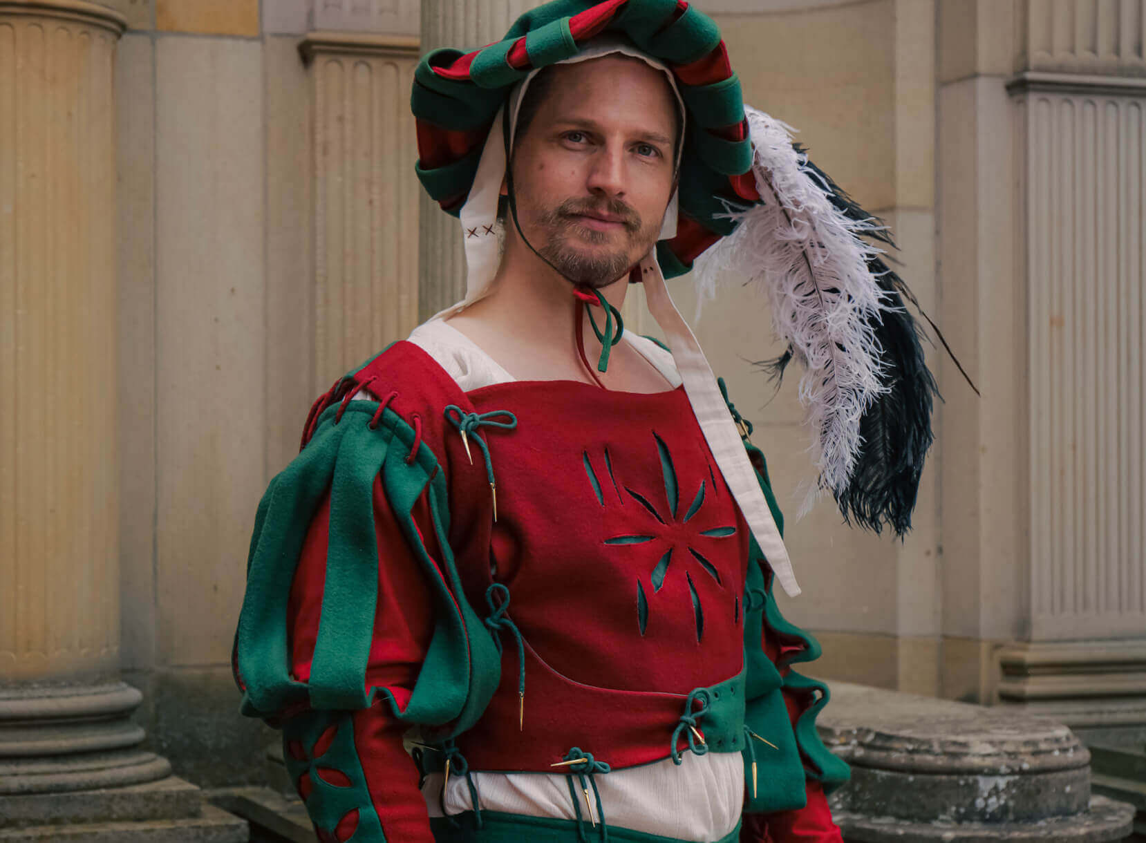 Man in traditional colorful medieval attire with a feathered hat, posing near classical architecture.