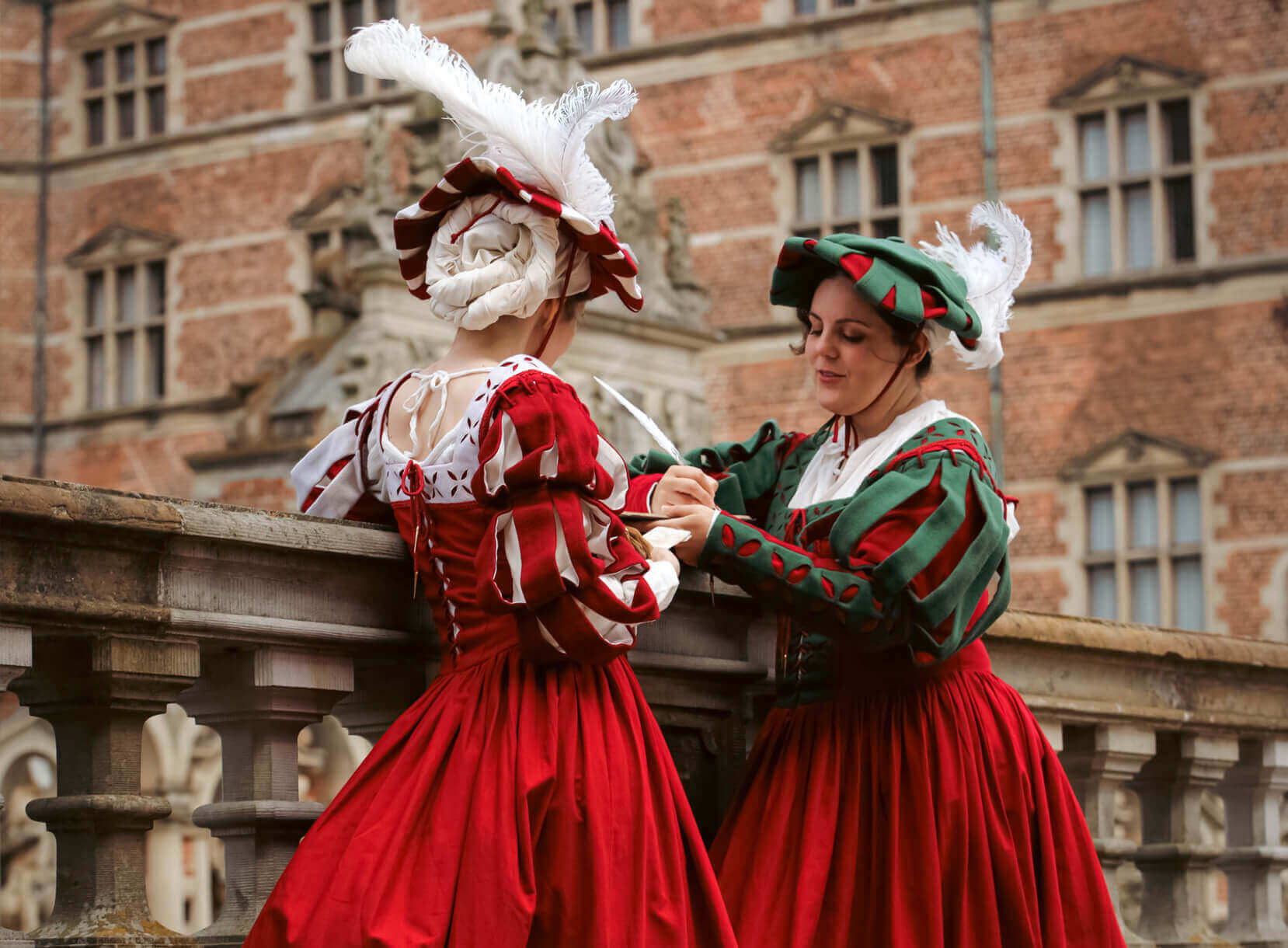 Two women in historical attire, wearing red dresses with white and green hats, conversing on a balcony.