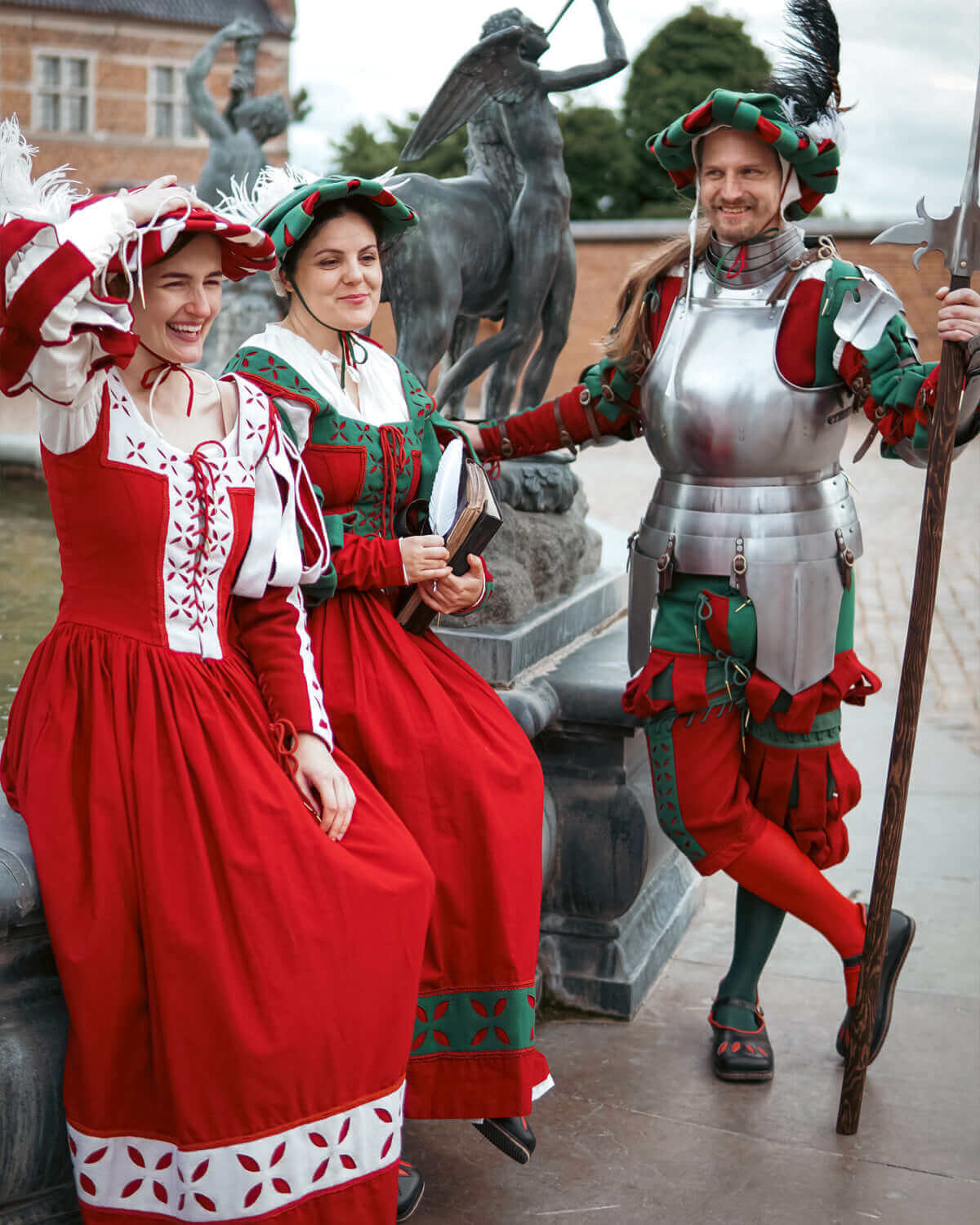 Three performers in medieval costumes: two women in red dresses and a man in armored attire, posing outdoors.