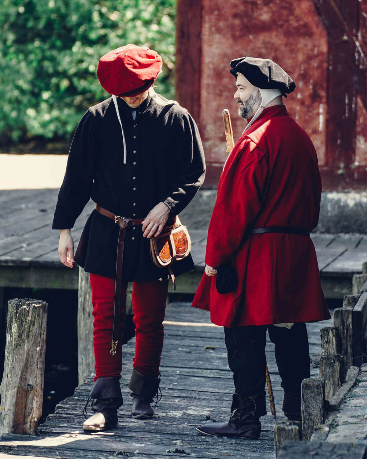 Two men in historical costumes walking on a wooden dock, showcasing 15th-century fashion with hats and colorful clothing.