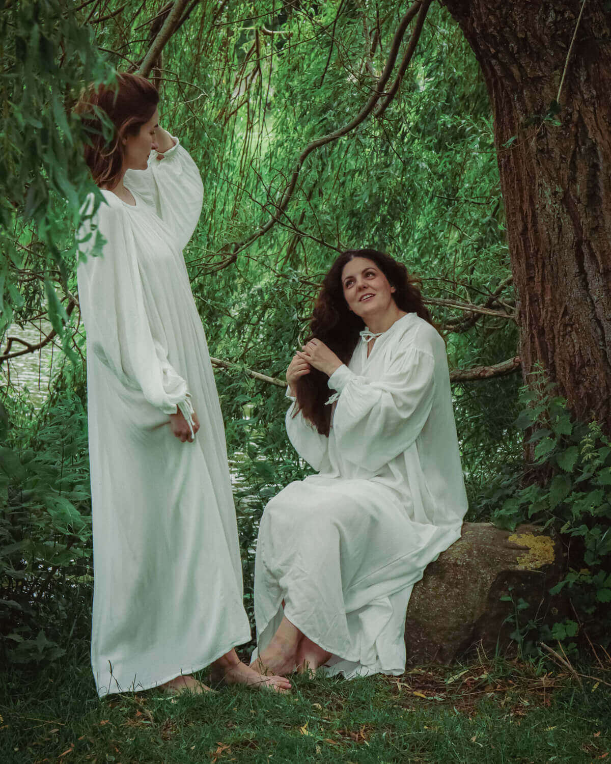 Two women in flowing white dresses sitting by a tree, one braiding the other's hair in a serene, green setting.