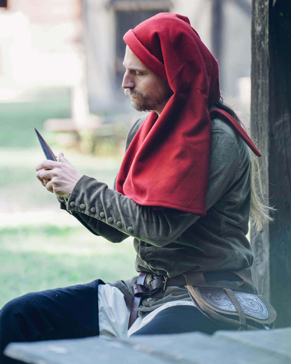 A man wearing a red hooded cloak sits outdoors, examining a knife, showcasing historical attire and craftsmanship.