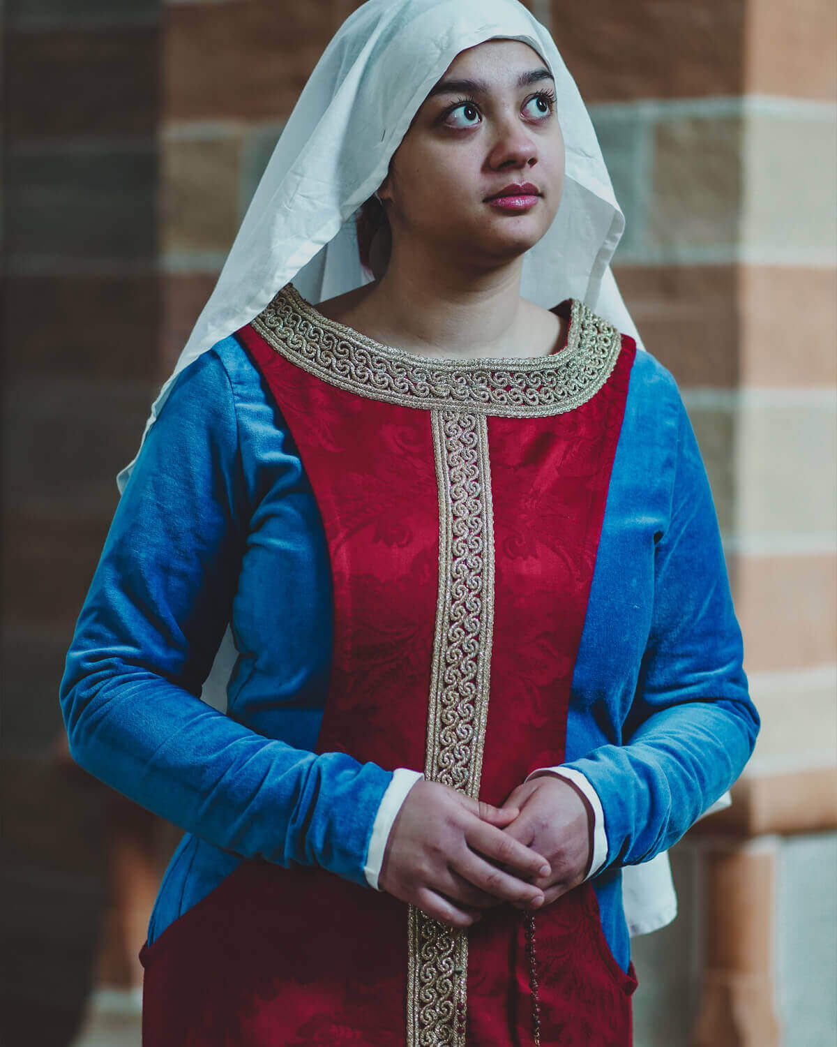 Woman in historical blue and red medieval gown with white veil, looking up.