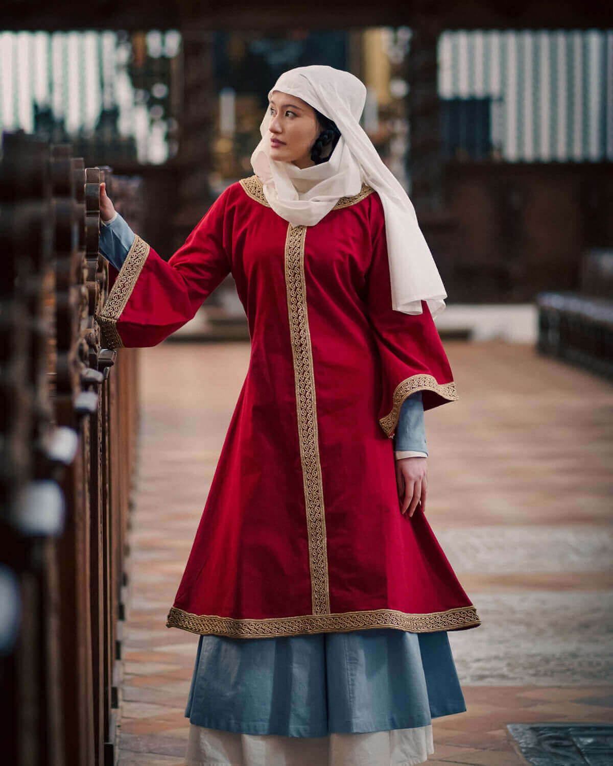 Woman in red medieval overtunic and white wimple, standing in a historical setting.